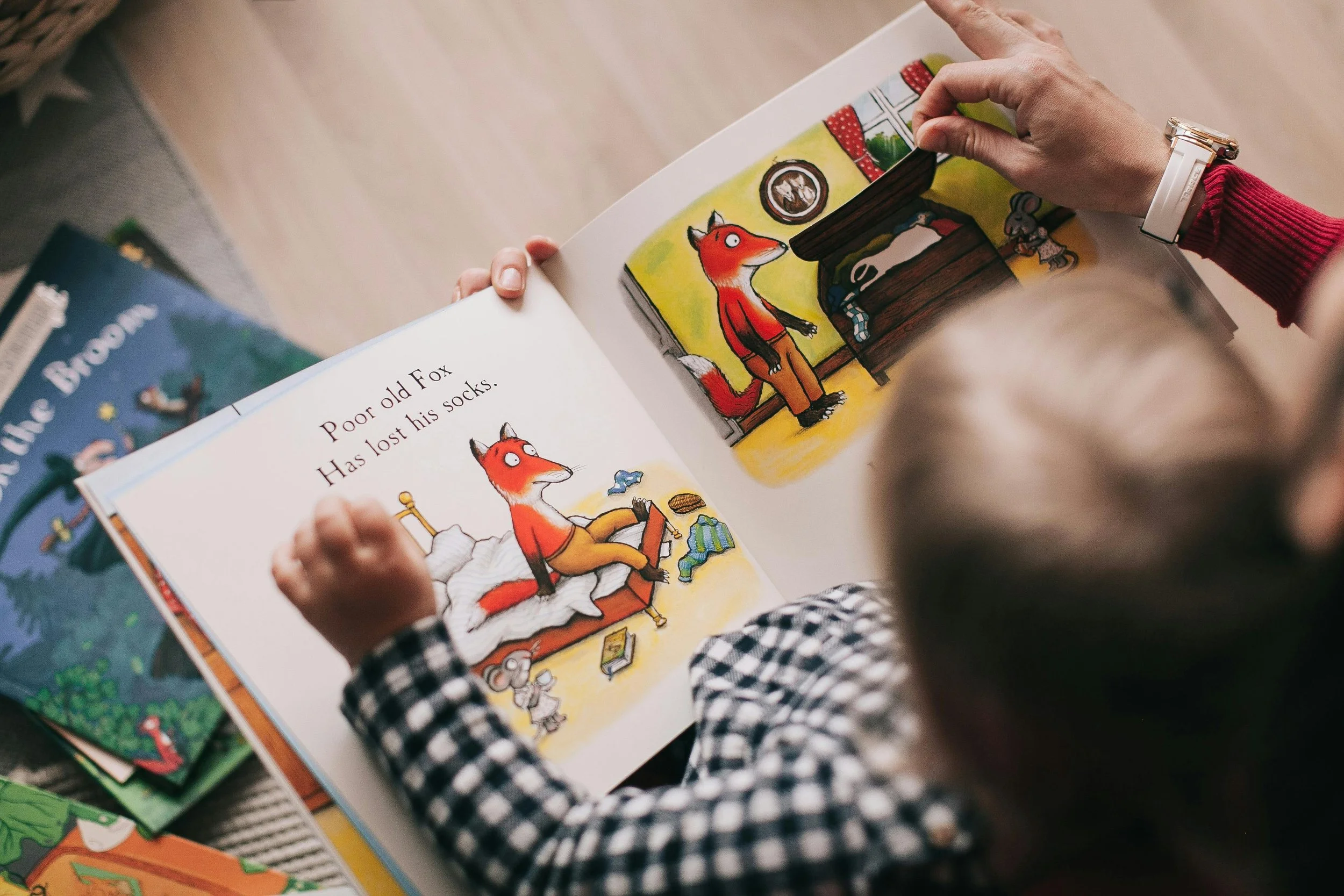 A person reading a children's book to a young child. The book shows an illustration of a fox missing his socks, with another fox standing in a room filled with furniture and toys.