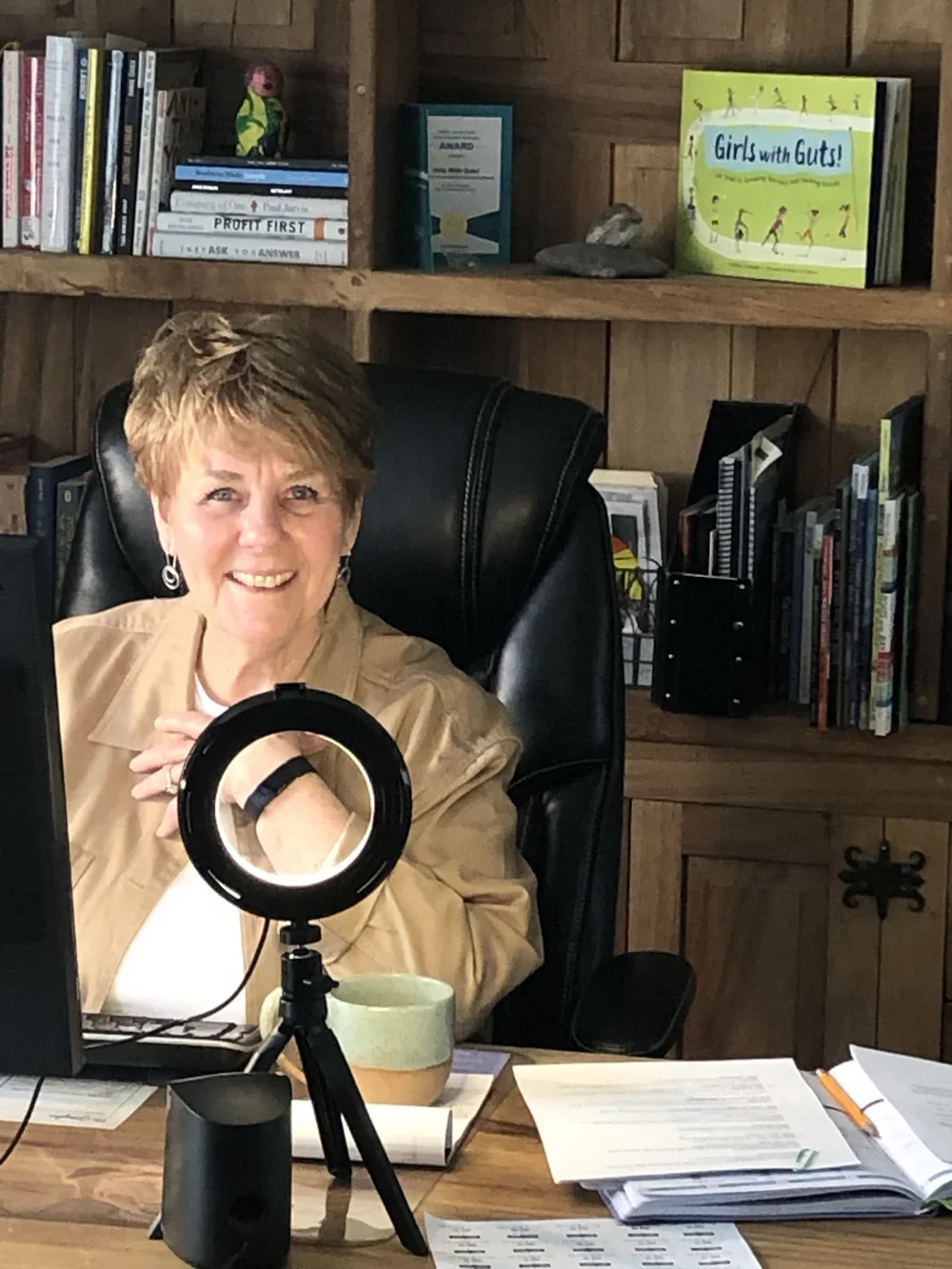 Debbie Gonzales, Founder, sitting at a wooden desk with a ring light, papers, and a mug. She is seated in a black leather office chair, with a wooden bookshelf behind her filled with books, including titles like "Girls with Guts!" and "Profit First."