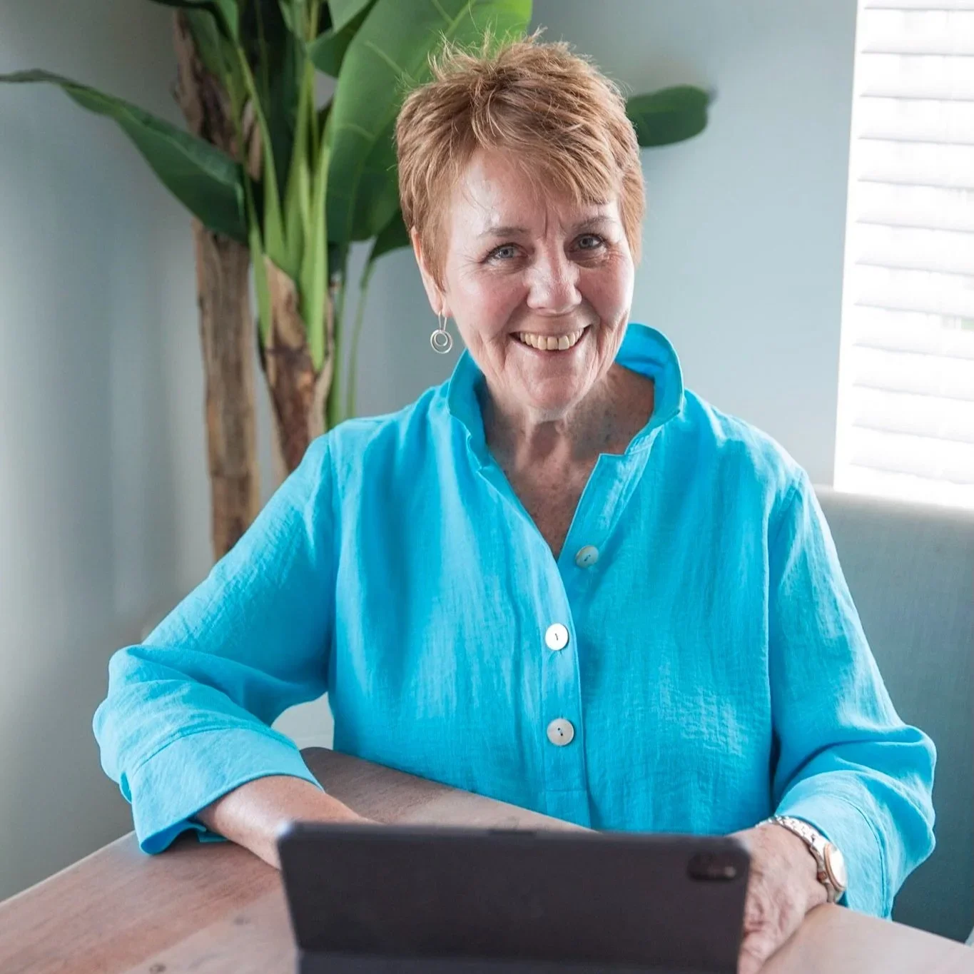 A smiling woman with short red hair wearing a light blue blouse sitting at a wooden table with a tablet device.