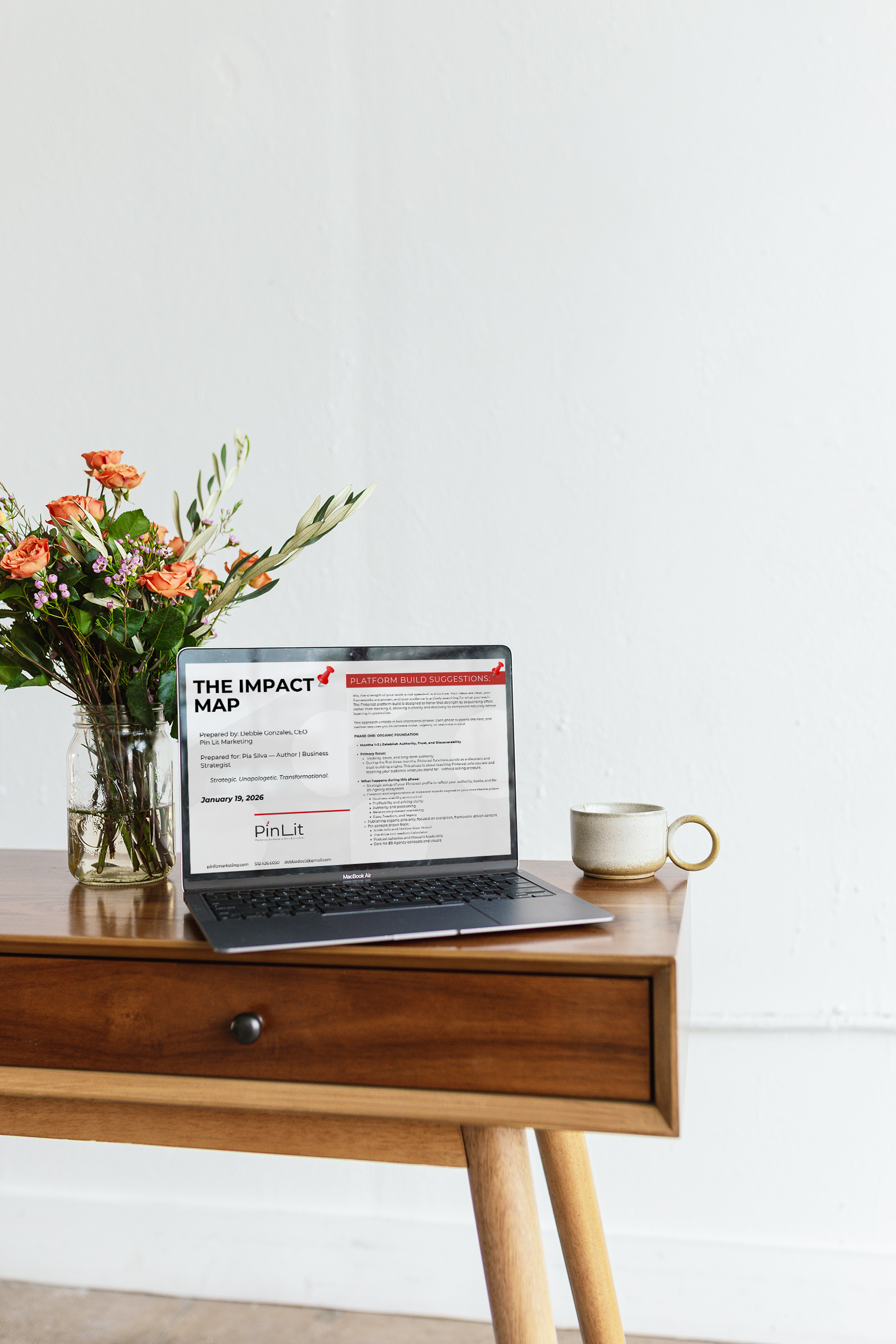 Wooden desk with an open MacBook Air displaying a presentation titled 'The Impact Map', a glass jar with a bouquet of pink and purple flowers, a beige coffee mug, and a plain white wall in the background.