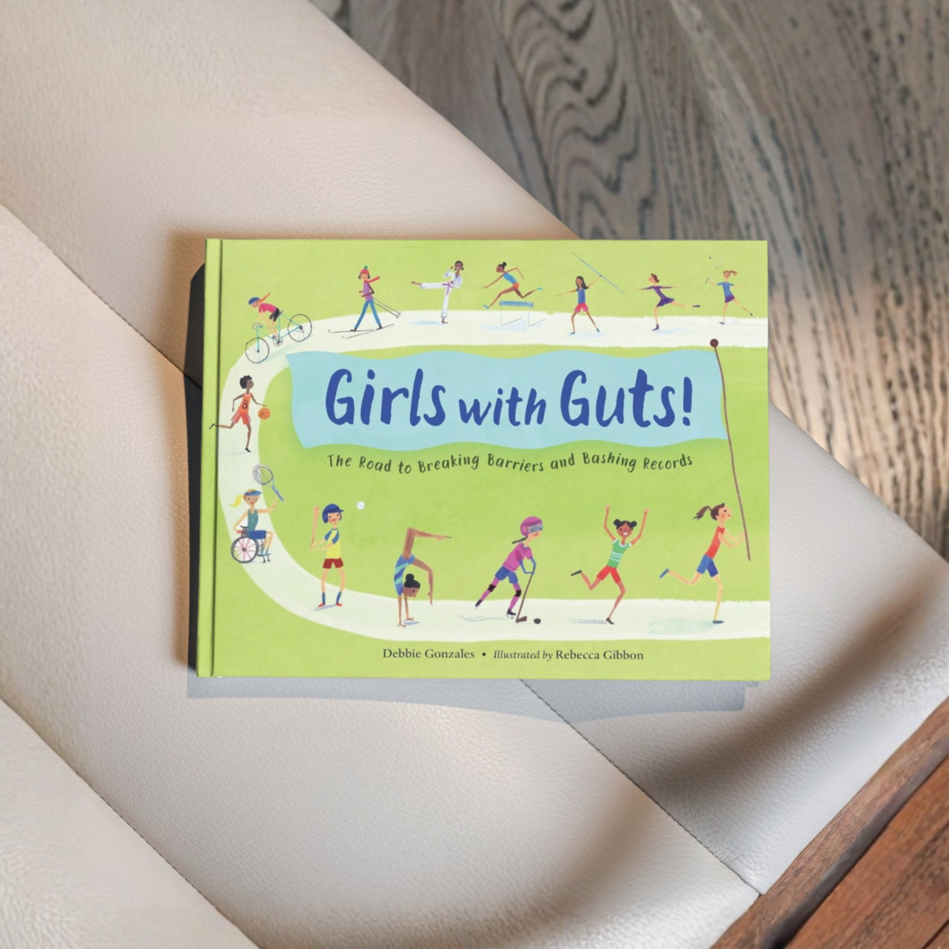 A book titled 'Girls with Guts! The Road to Breaking Barriers and Bashing Records' on a light-colored chair, featuring colorful illustrations of girls participating in various sports on its cover.