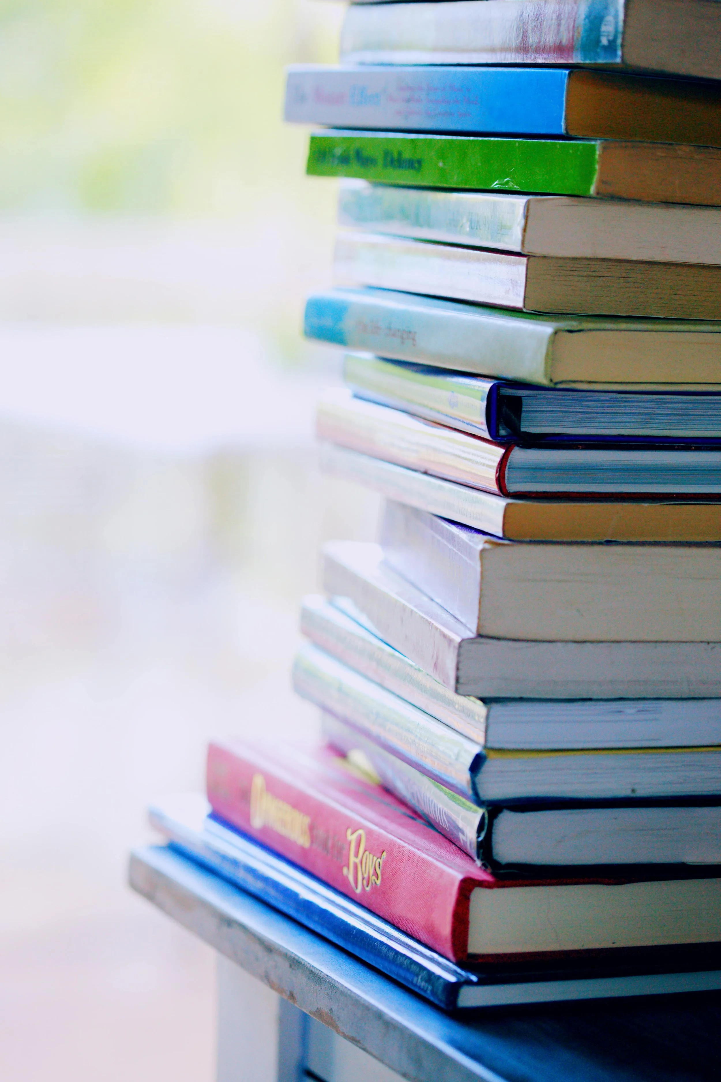 Stack of colorful books on a table near a window with blurred outdoor view.