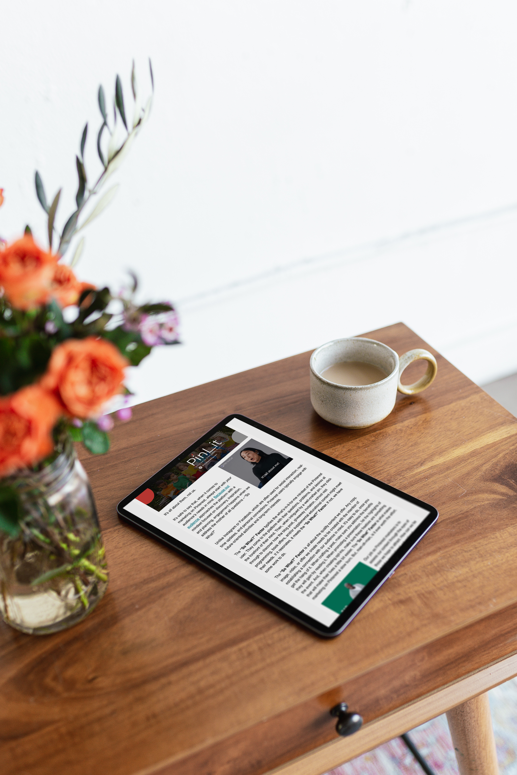 A wooden table with a tablet displaying an article, a ceramic mug filled with a beverage, and a vase of colorful flowers.