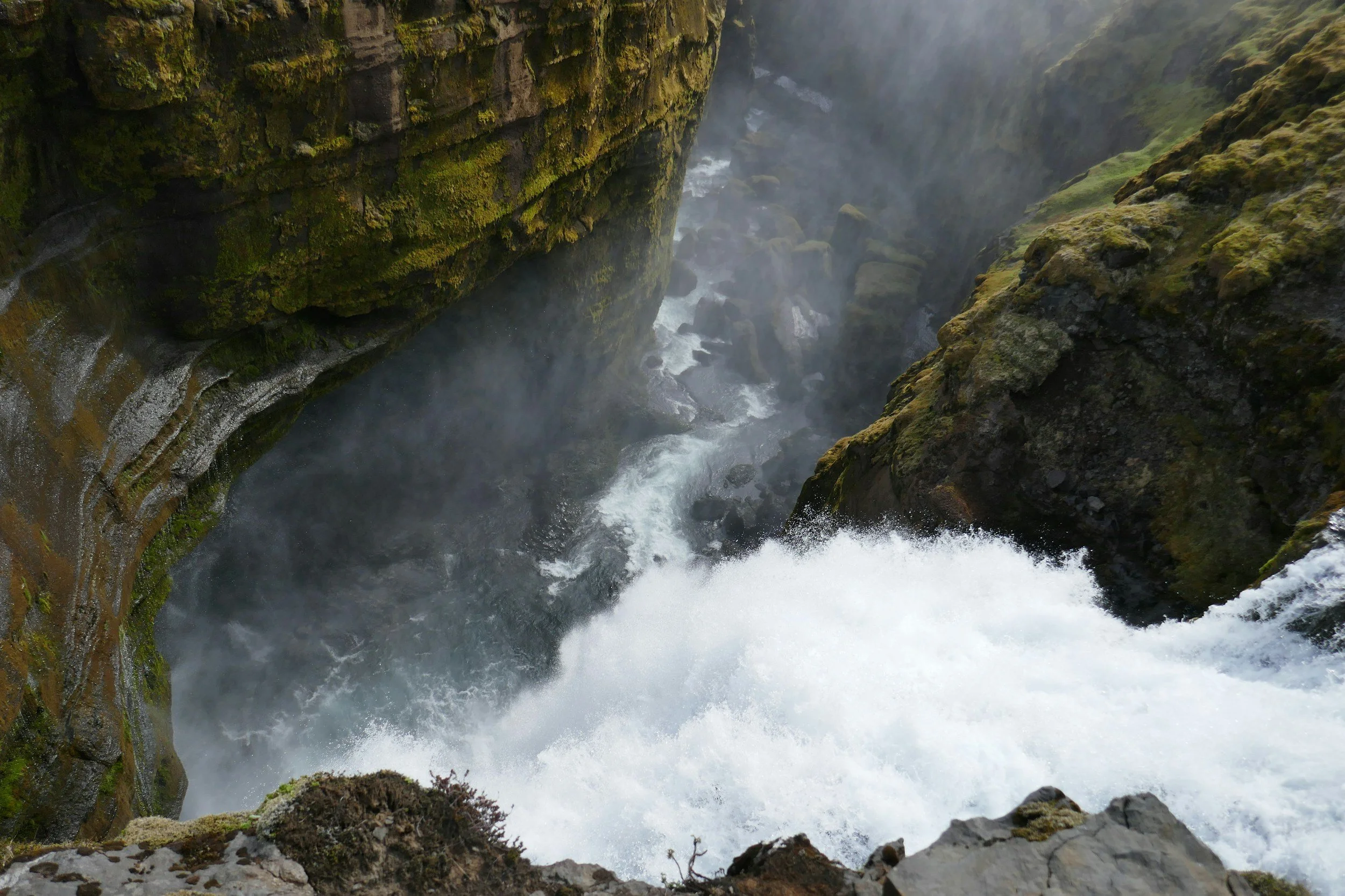 A rushing river flowing through a deep, moss-covered canyon with steep rocky walls and mist rising from the water.