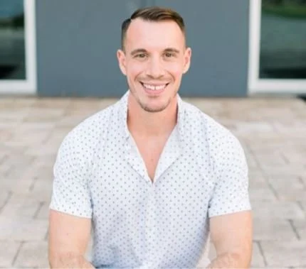 A smiling man in a white, short-sleeved, polka dot shirt sitting outdoors in front of a gray building with windows.