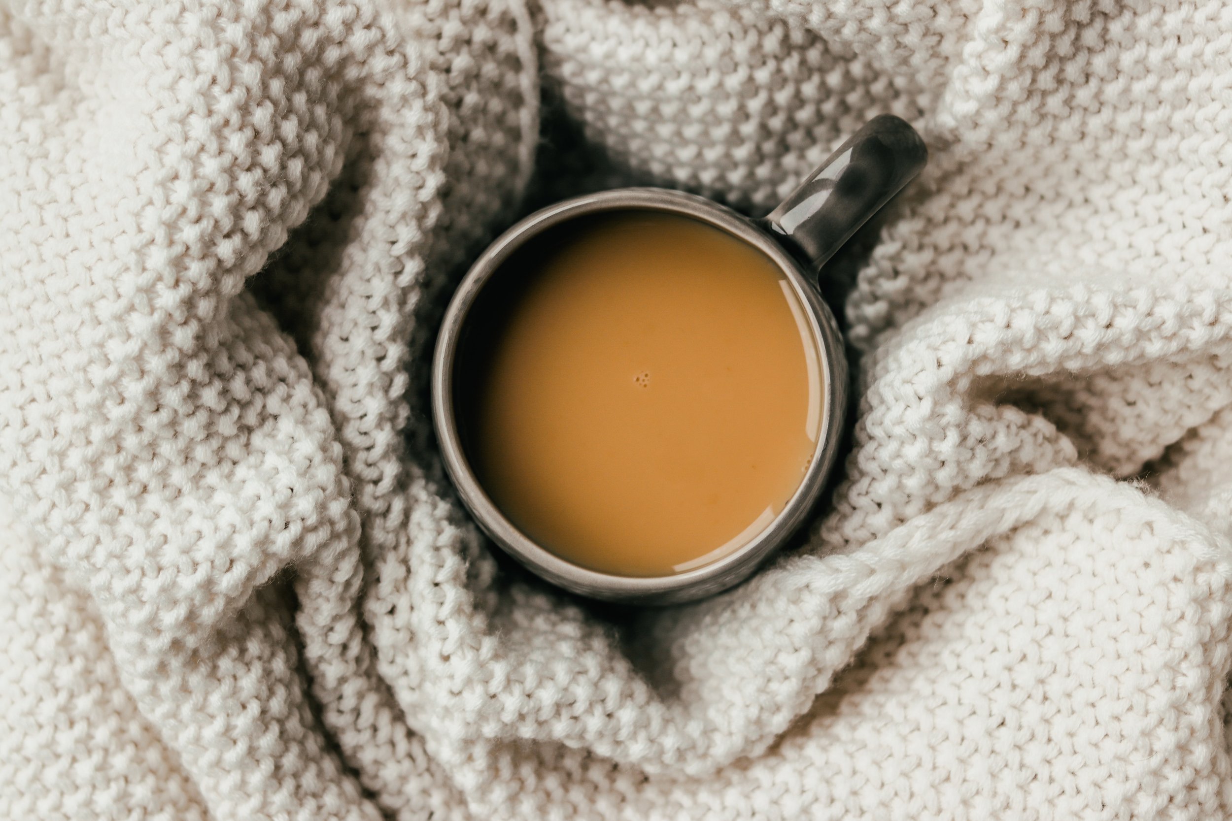 A top-down view of a mug filled with tea or coffee, surrounded by a cozy, chunky, cream-colored knit blanket.