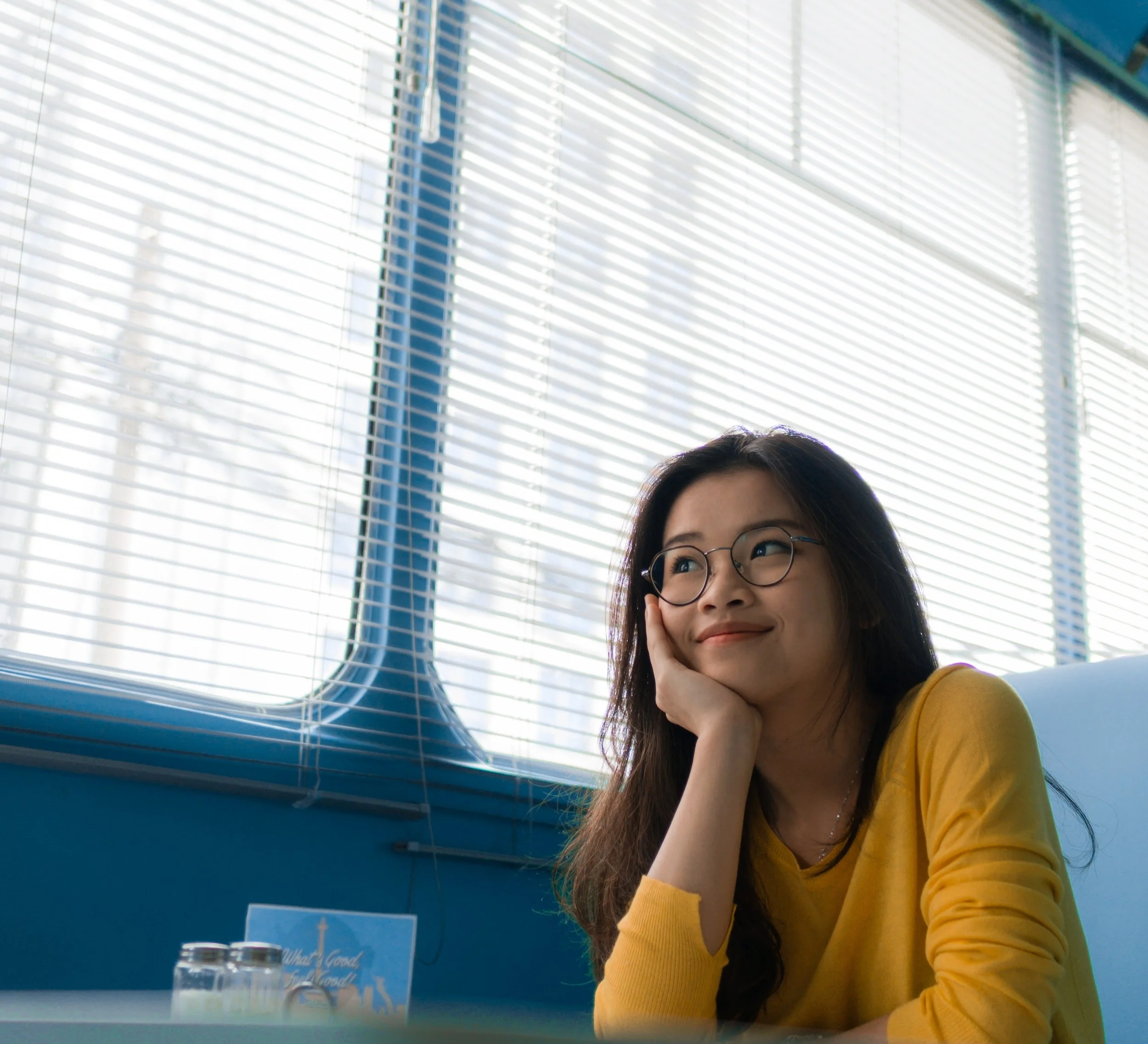 Young woman with glasses and a yellow shirt sitting at a table, resting her head on her hand, smiling and looking to the side, with blinds and a window in the background.