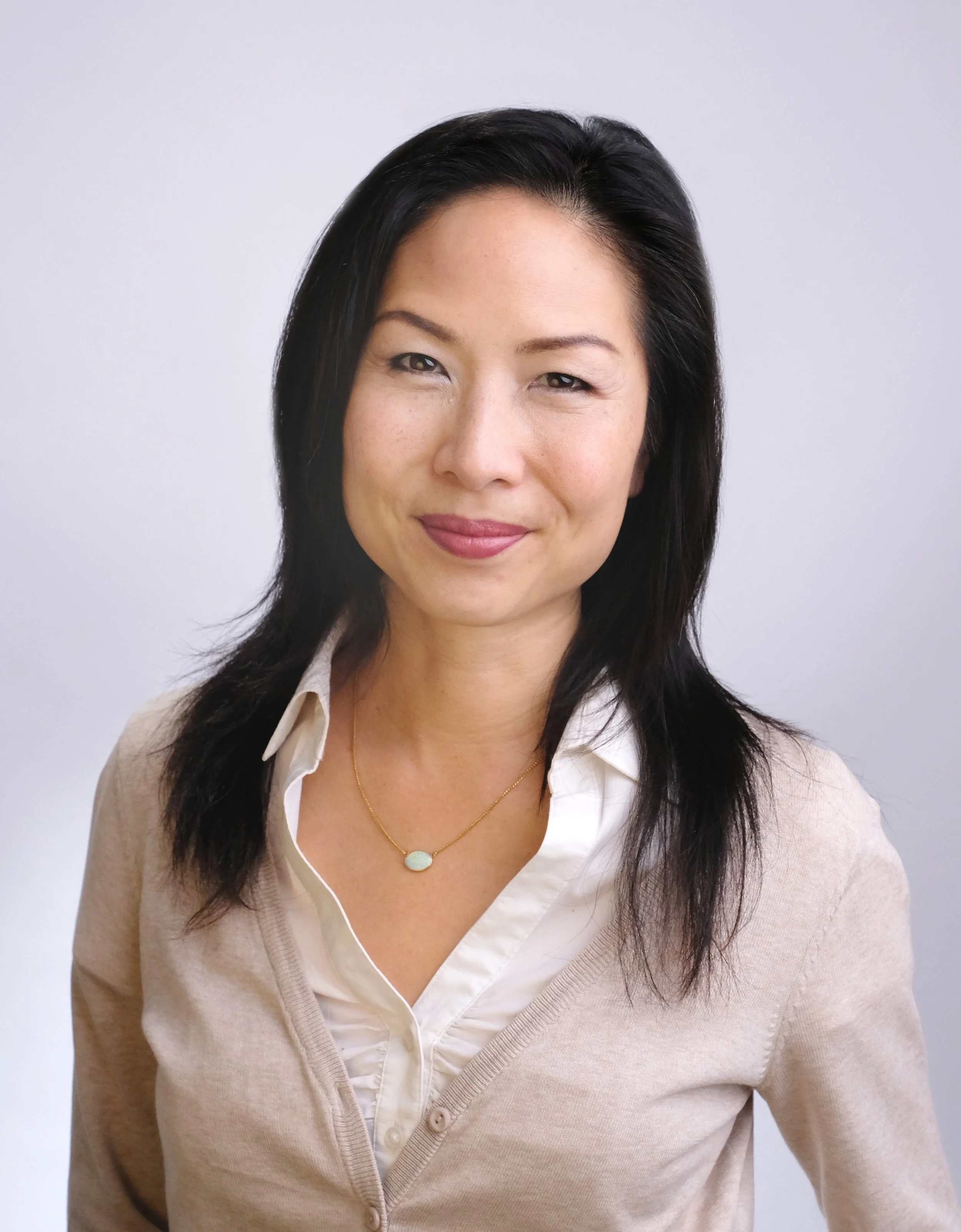A woman with black hair wearing a white blouse, a beige cardigan, and a gold necklace, smiling in front of a plain light gray background.