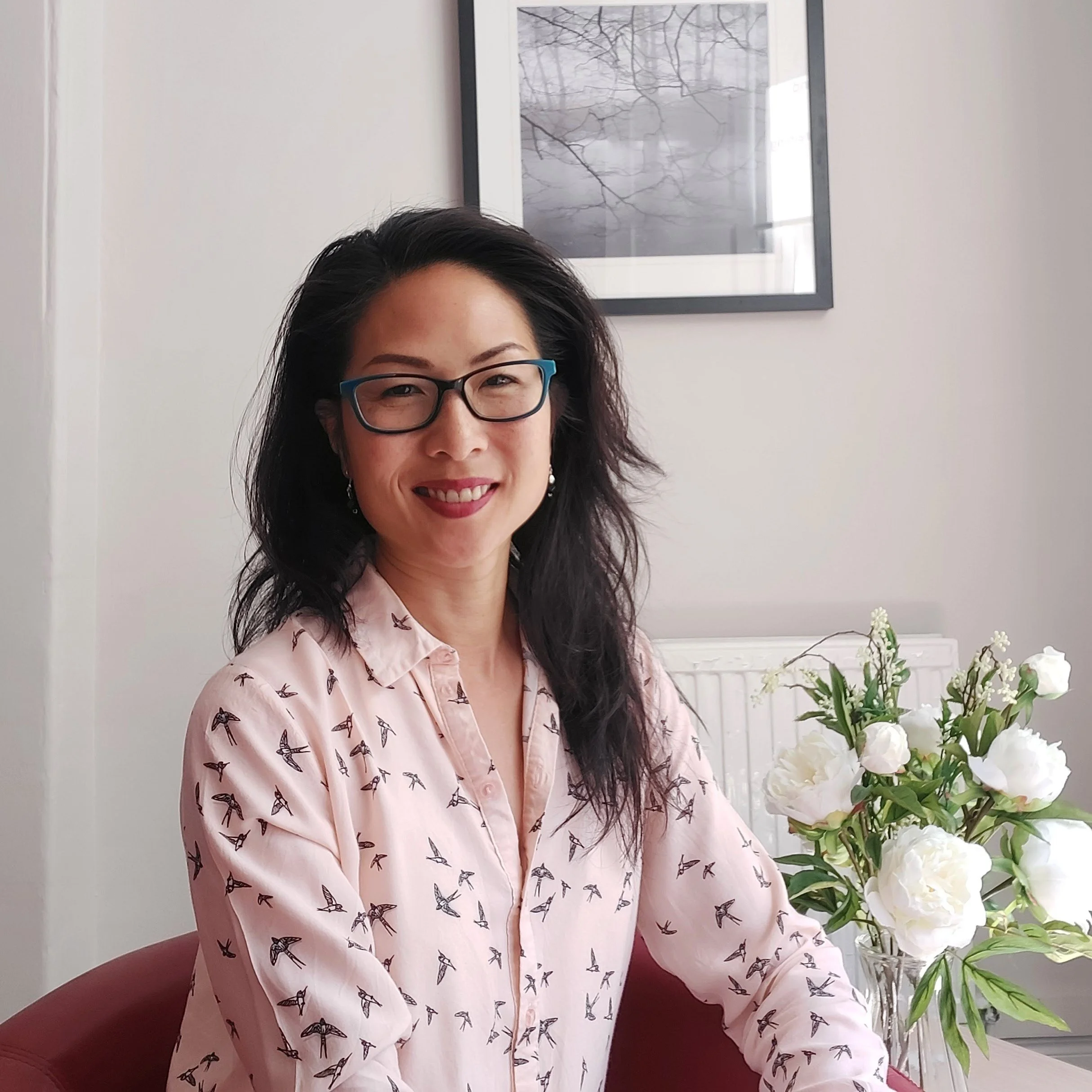 A smiling woman with black wavy hair, glasses with blue frames, wearing a light pink button-up shirt with a bird print, sitting at a table next to a vase of white flowers, with a picture of tree branches on the wall in the background.