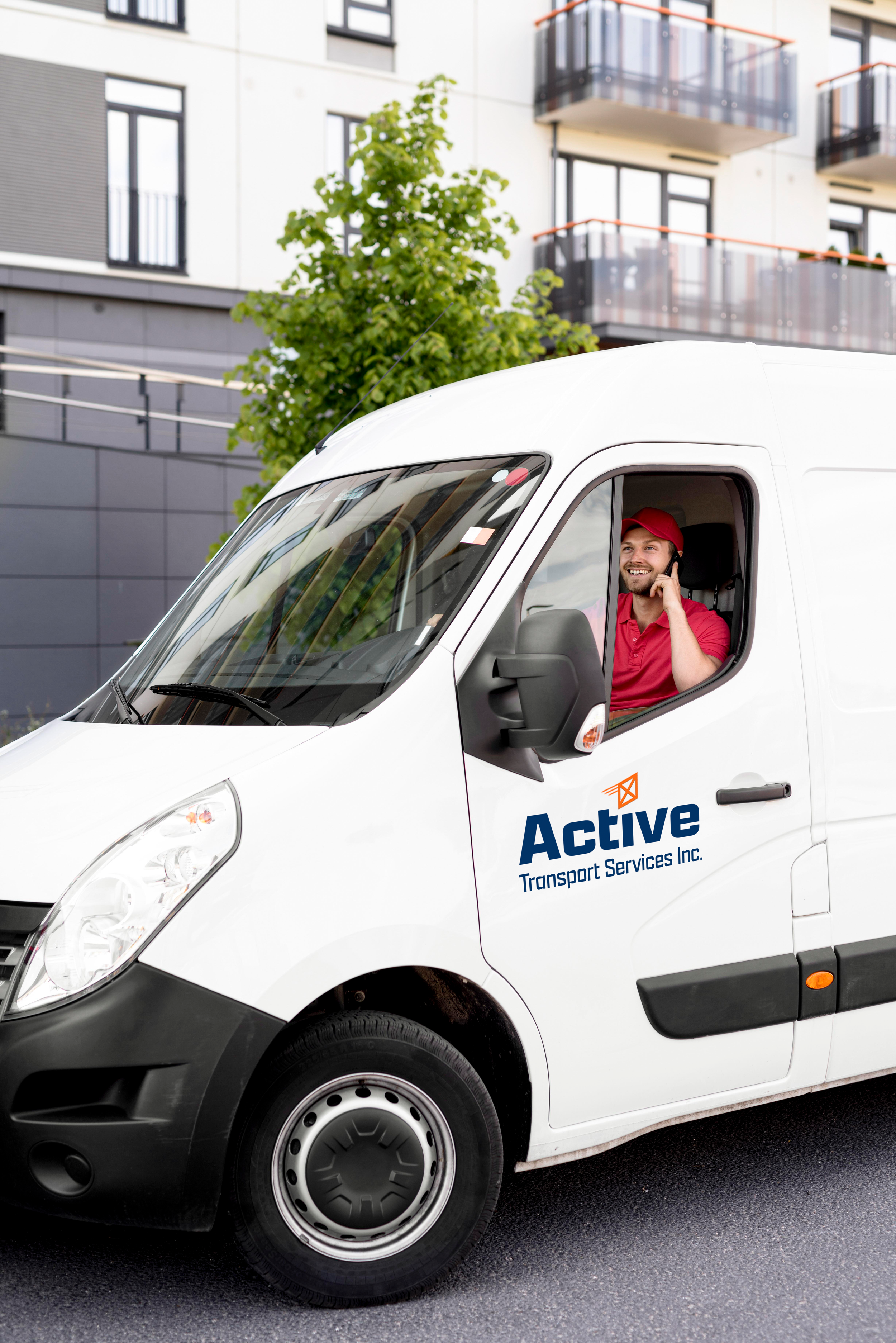 A delivery driver in a red cap and shirt is sitting in the driver's seat of a white delivery van marked 'Active Transport Services Inc.' The van is parked on a city street with modern apartment buildings and a tree in the background.