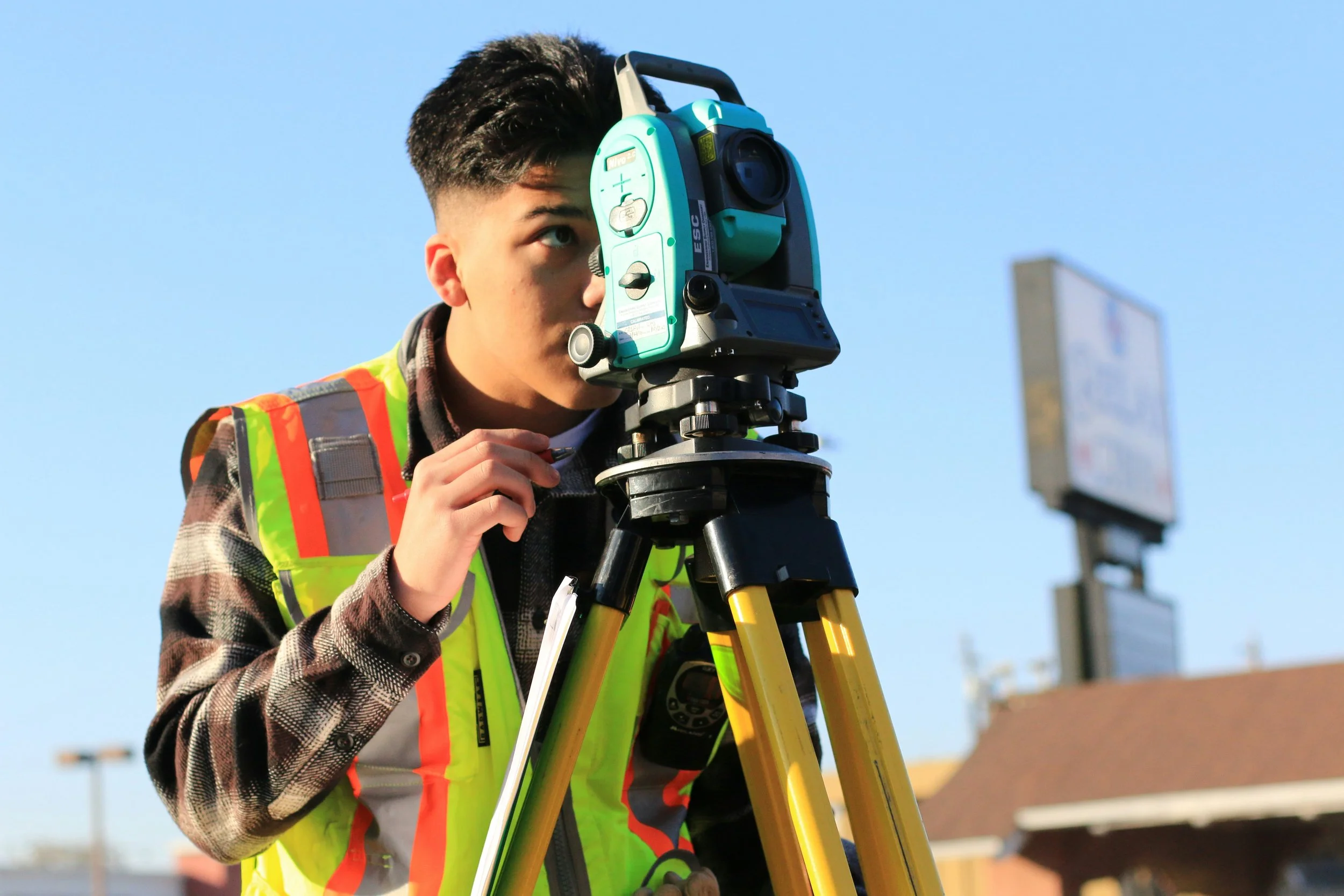 A surveyor is looking through a theodolite mounted on a yellow tripod, wearing a high-visibility vest, outdoors in front of a clear blue sky.