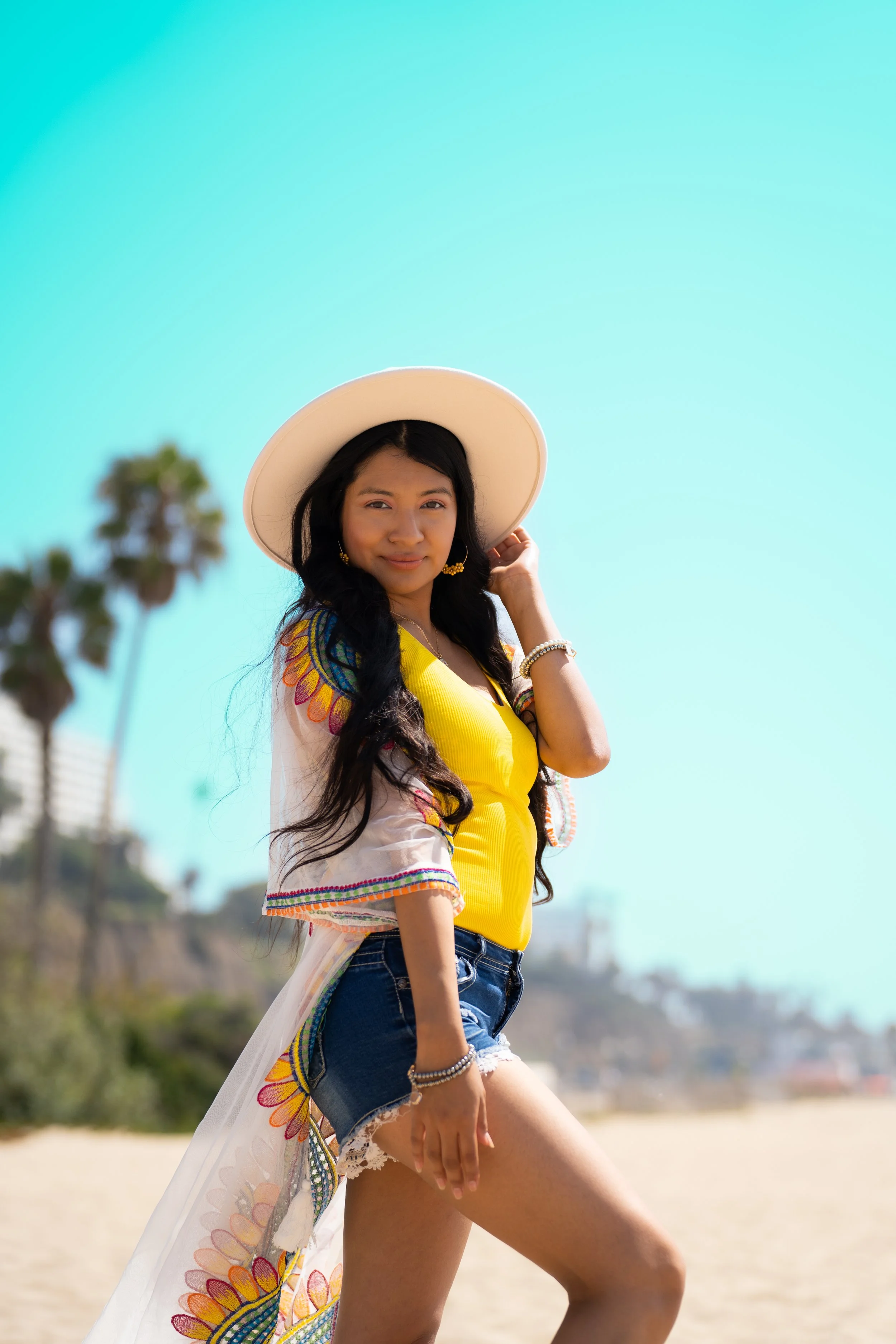 Young woman in a yellow top and denim shorts at the beach, wearing a large sun hat and jewelry, with palm trees and a clear blue sky in the background.