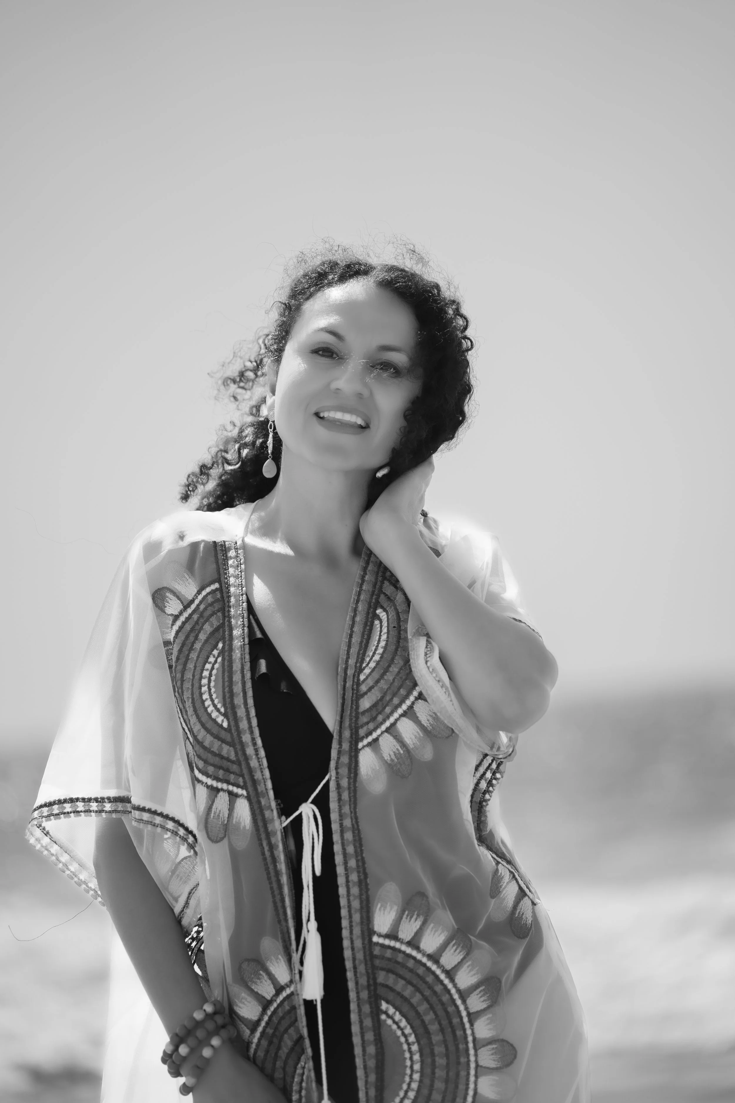 Black and white photo of a woman with curly hair wearing a patterned cover-up, smiling, and standing outdoors.