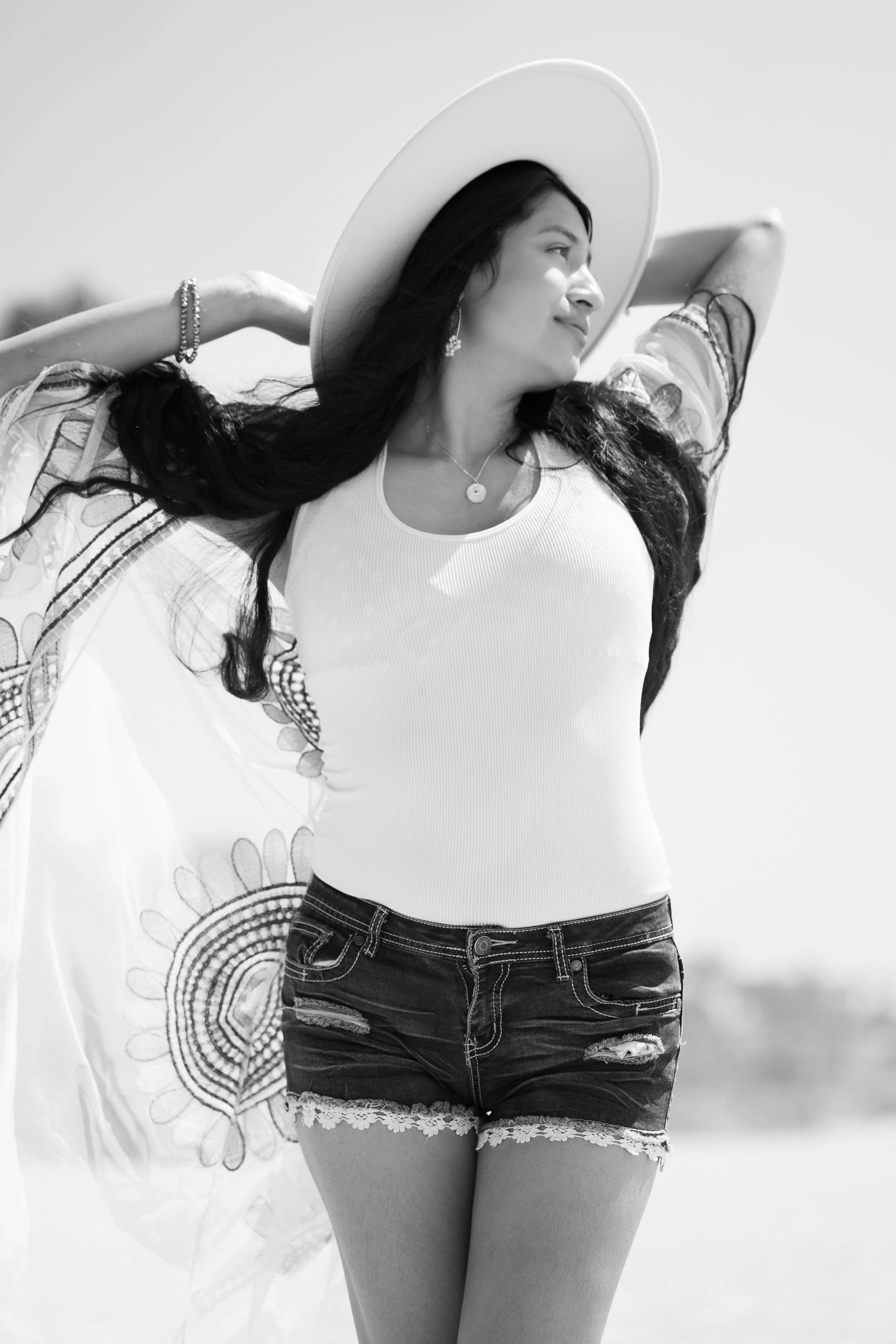 Woman with long dark hair wearing a wide-brimmed hat, white tank top, and denim shorts with lace trim, smiling outdoors in bright sunlight.