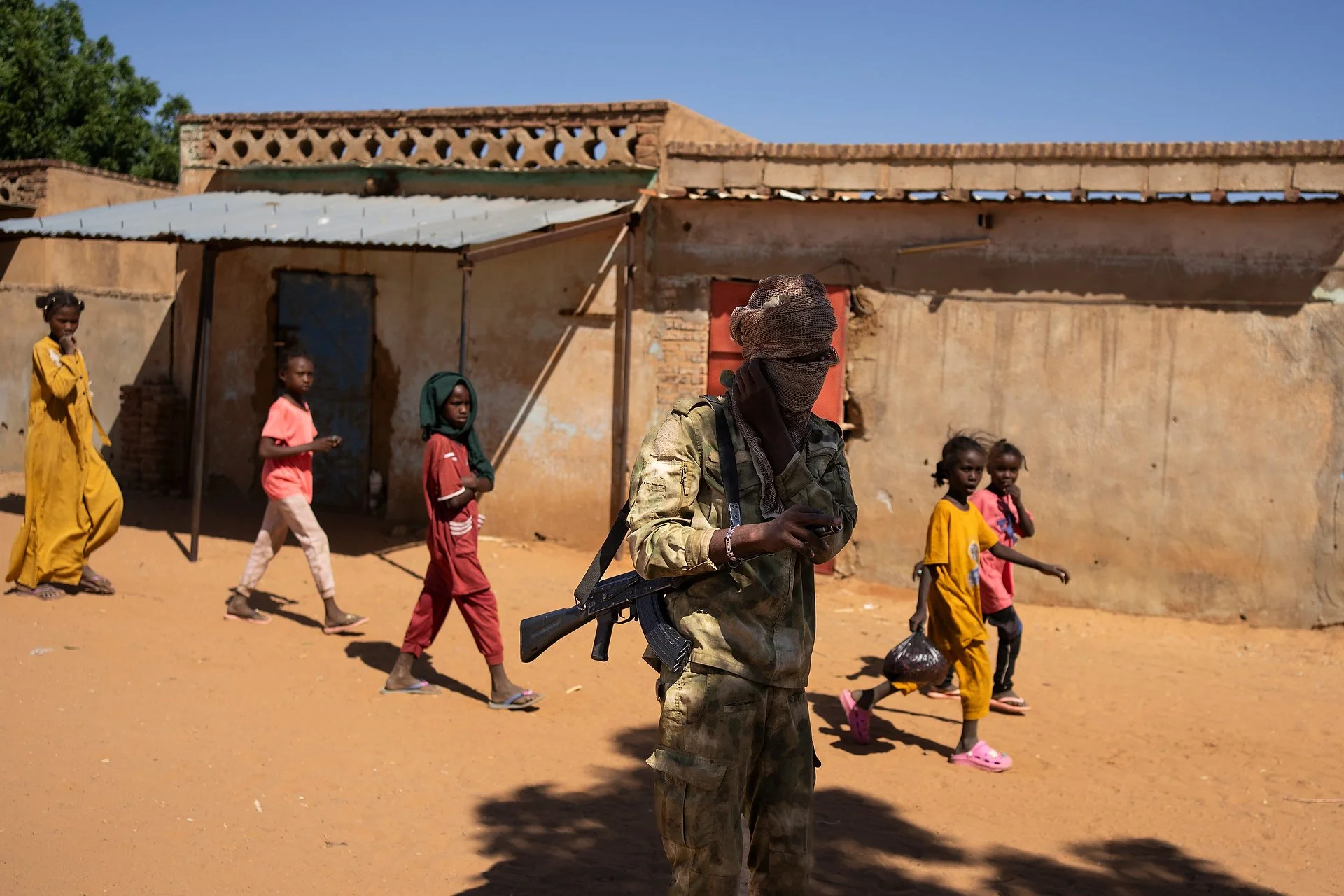 Man carrying a machine gun stands in the street in Sudan with children walking behind him.
