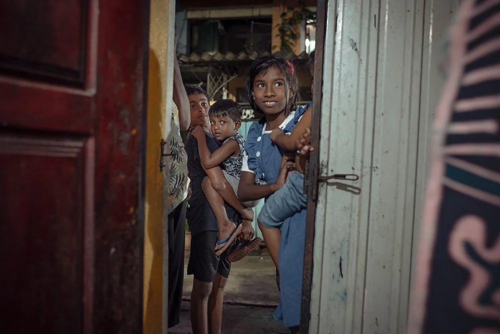 A young girl and boy holding younger children in their arms look out of an open door in a home.