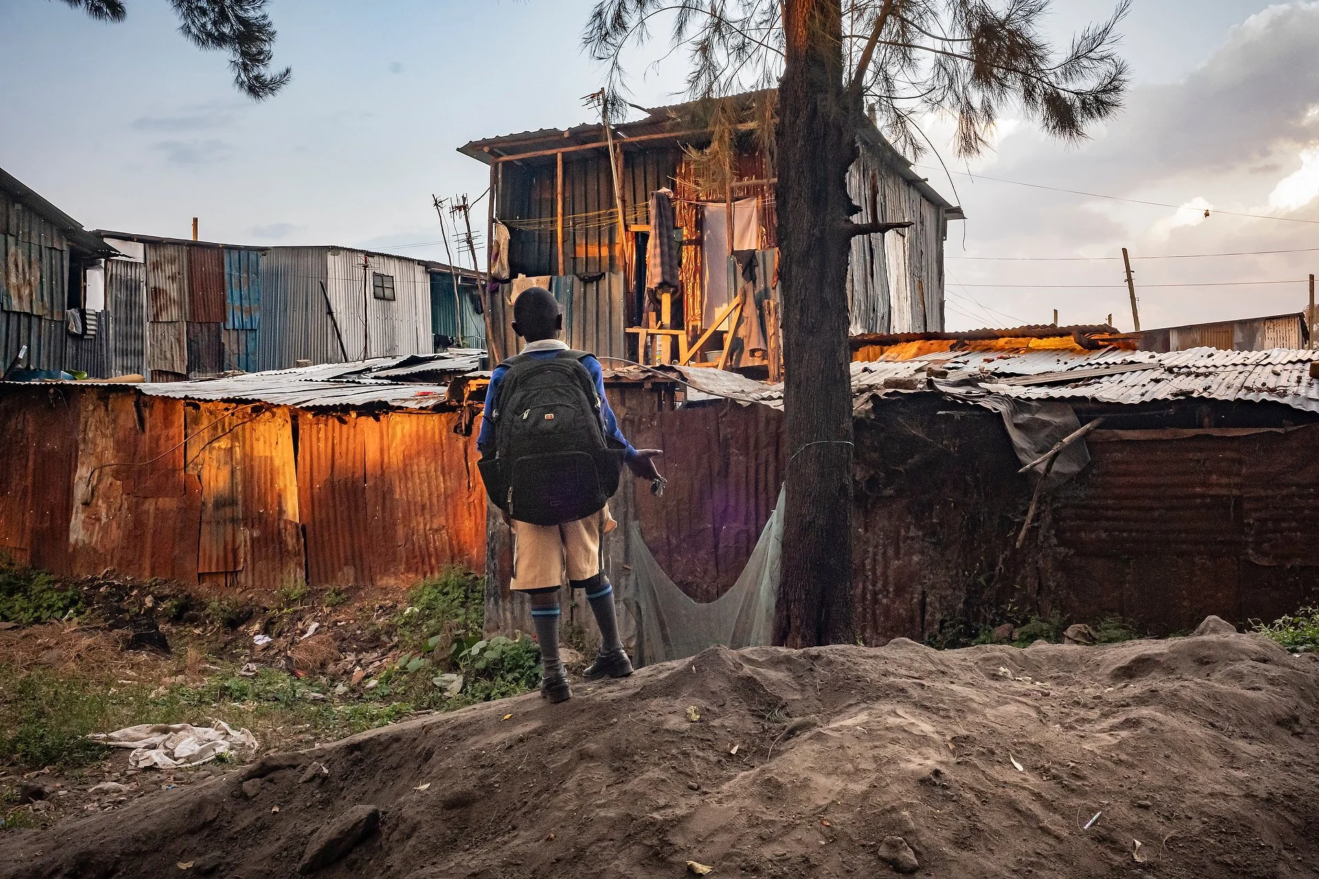 A child carrying a backpack stands on a mound of earth as he calls out to his mother at the school gate amid the bustle of Kibera, Nairobi, Kenya.