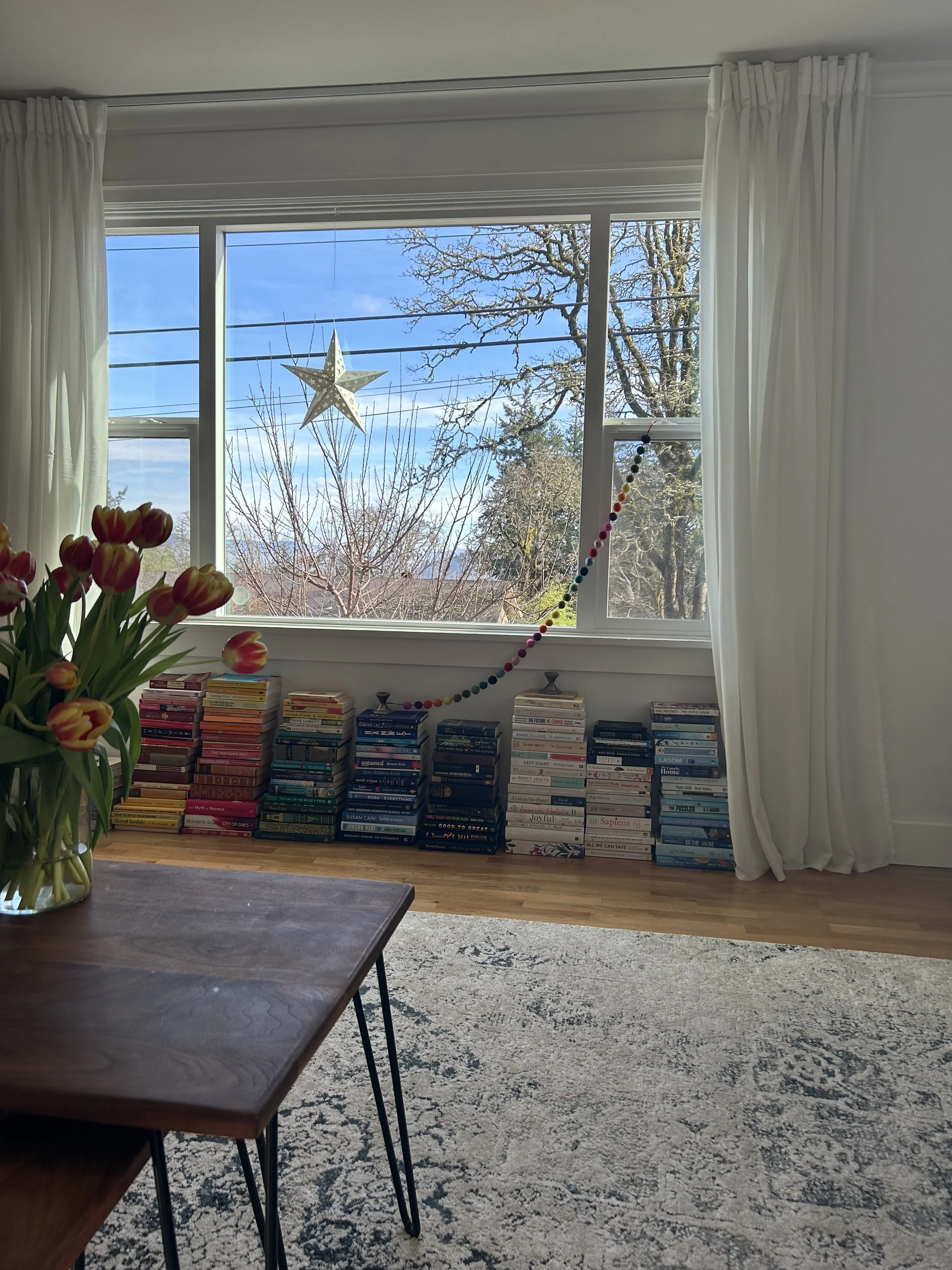 A living room window with white curtains, overlooking trees outside and decorated with a star and a colorful bead garland. Below, there are stacks of books arranged on the windowsill. In the foreground, a wooden table with a vase of tulips is partially visible, and a patterned rug covers the floor.