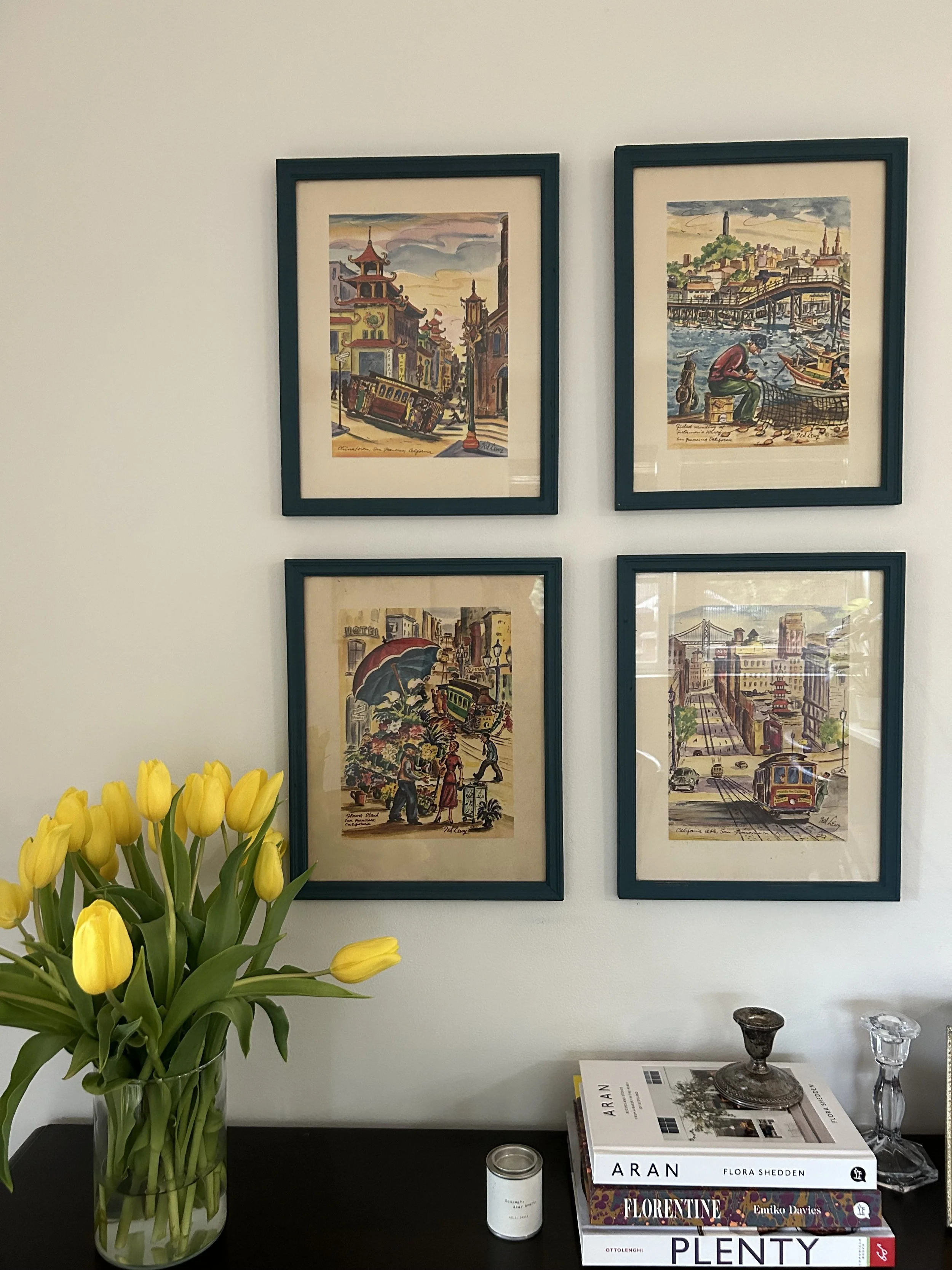 Four framed colorful cityscape and street scenes artwork hanging on a white wall.