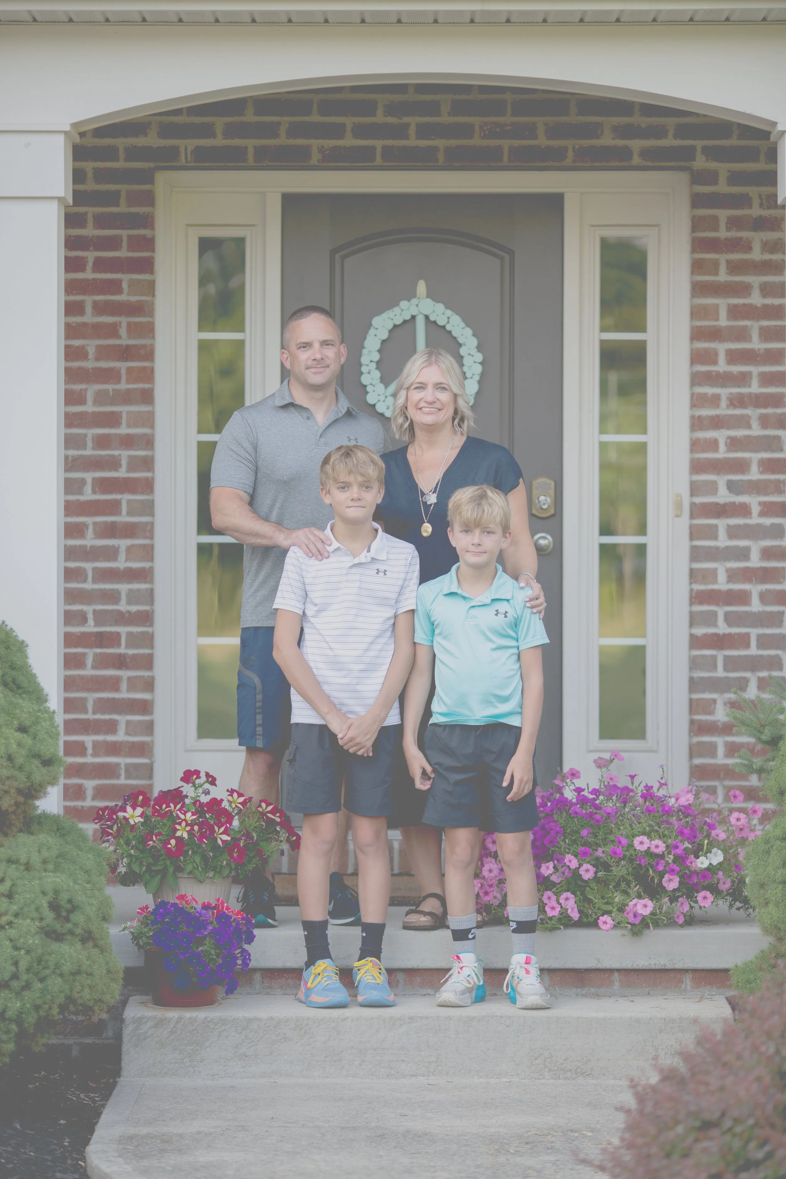 A family of four standing on the front porch of their brick house, smiling for the camera. The father and mother stand behind their two young sons in casual clothing. There are colorful flowers in pots on the porch, and a decorative wreath hangs on the front door.