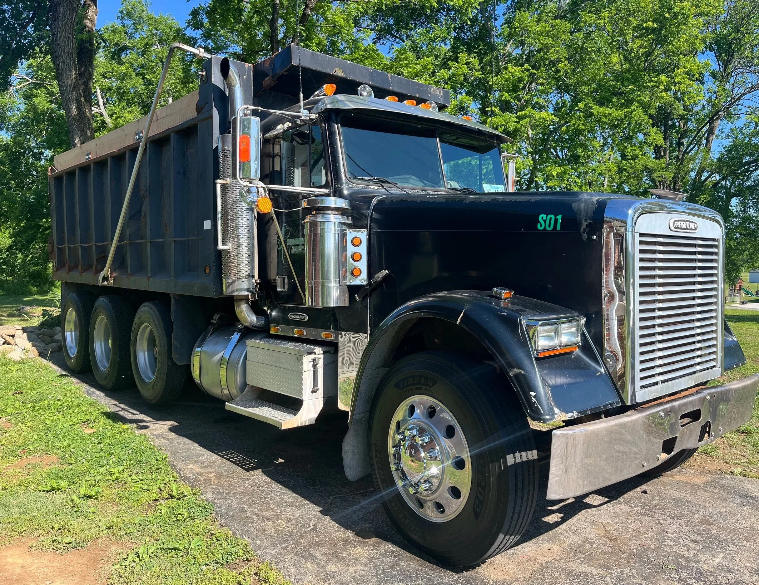 A black freightliner truck with a large grille and chrome details parked on a driveway with grass and trees in the background.