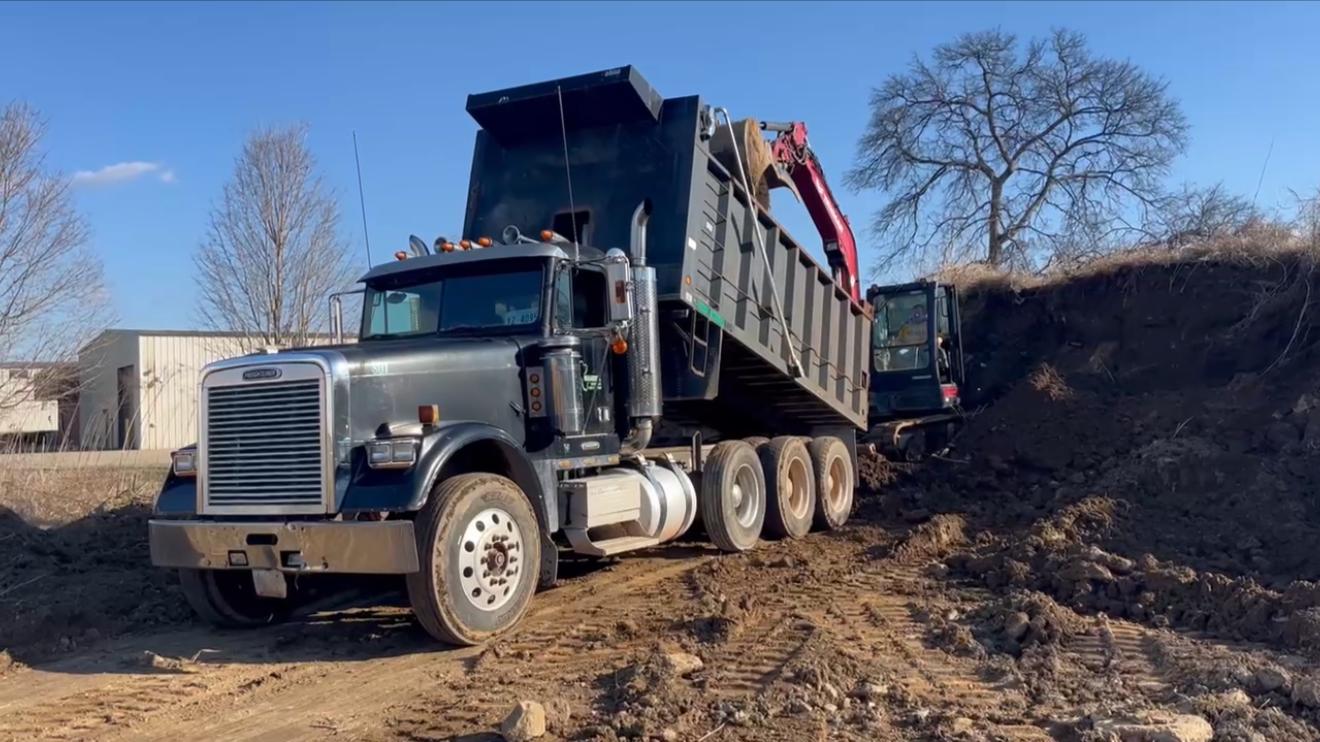 A large black dump truck and a smaller excavator working on a construction site with dirt and soil, trees in the background, and a clear blue sky.