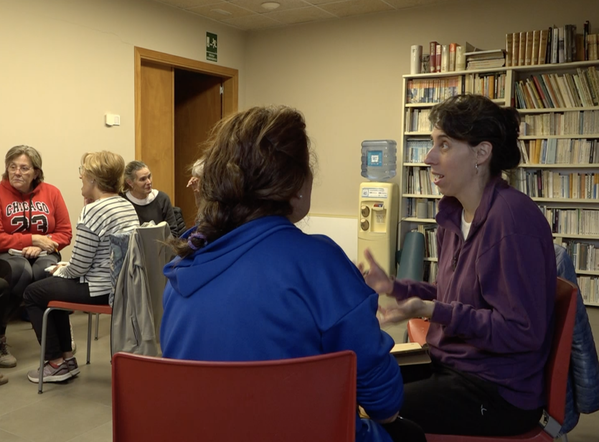 Women engaging in conversation in a room with bookshelves and reading material, while others sit in chairs in the background.