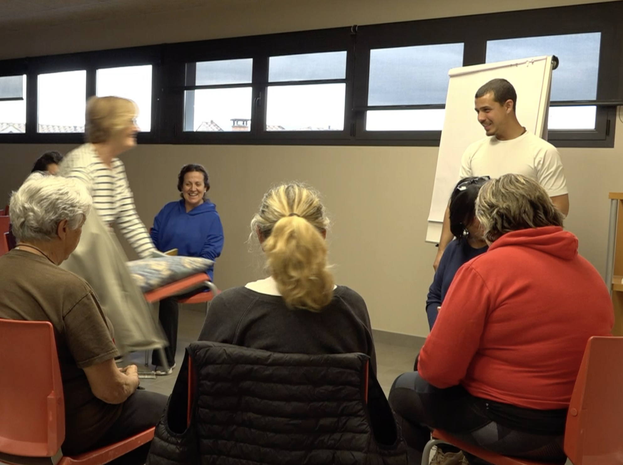A group of people gathered in a room, some sitting and some standing, engaged in a conversation or event. There is a man standing near a whiteboard, smiling, and women sitting in the audience. The room has windows on the upper part of the wall.