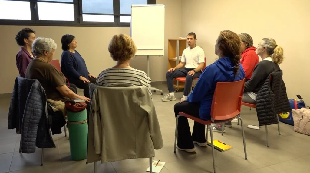 A group of people sitting in a semi-circle in a classroom or meeting room, listening to a man speaking. There is a whiteboard behind him and windows above.