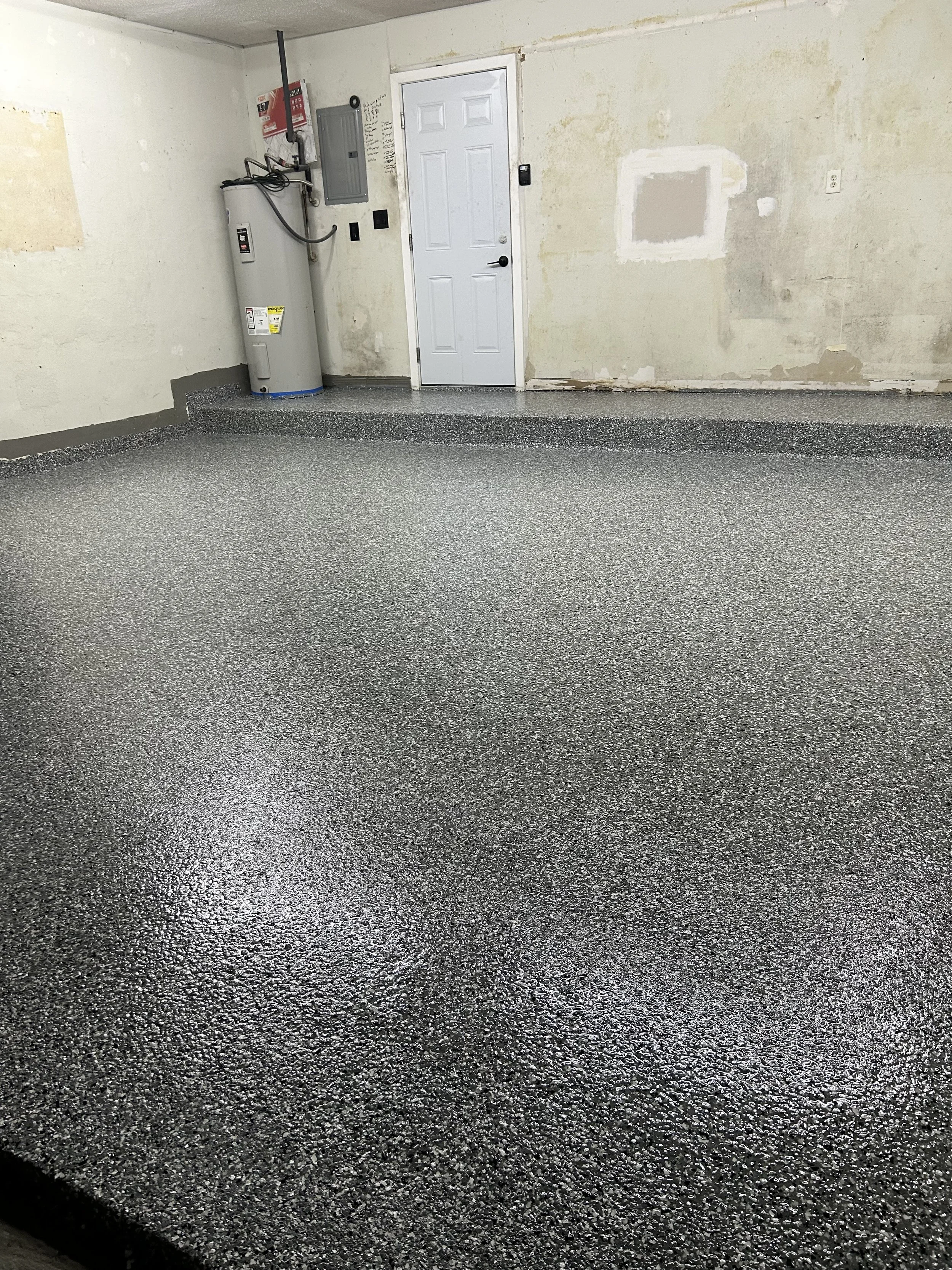 Empty garage with a textured gray epoxy-coated floor, unfinished walls, and a white door. A water heater and electrical panel are mounted on the wall near the door.