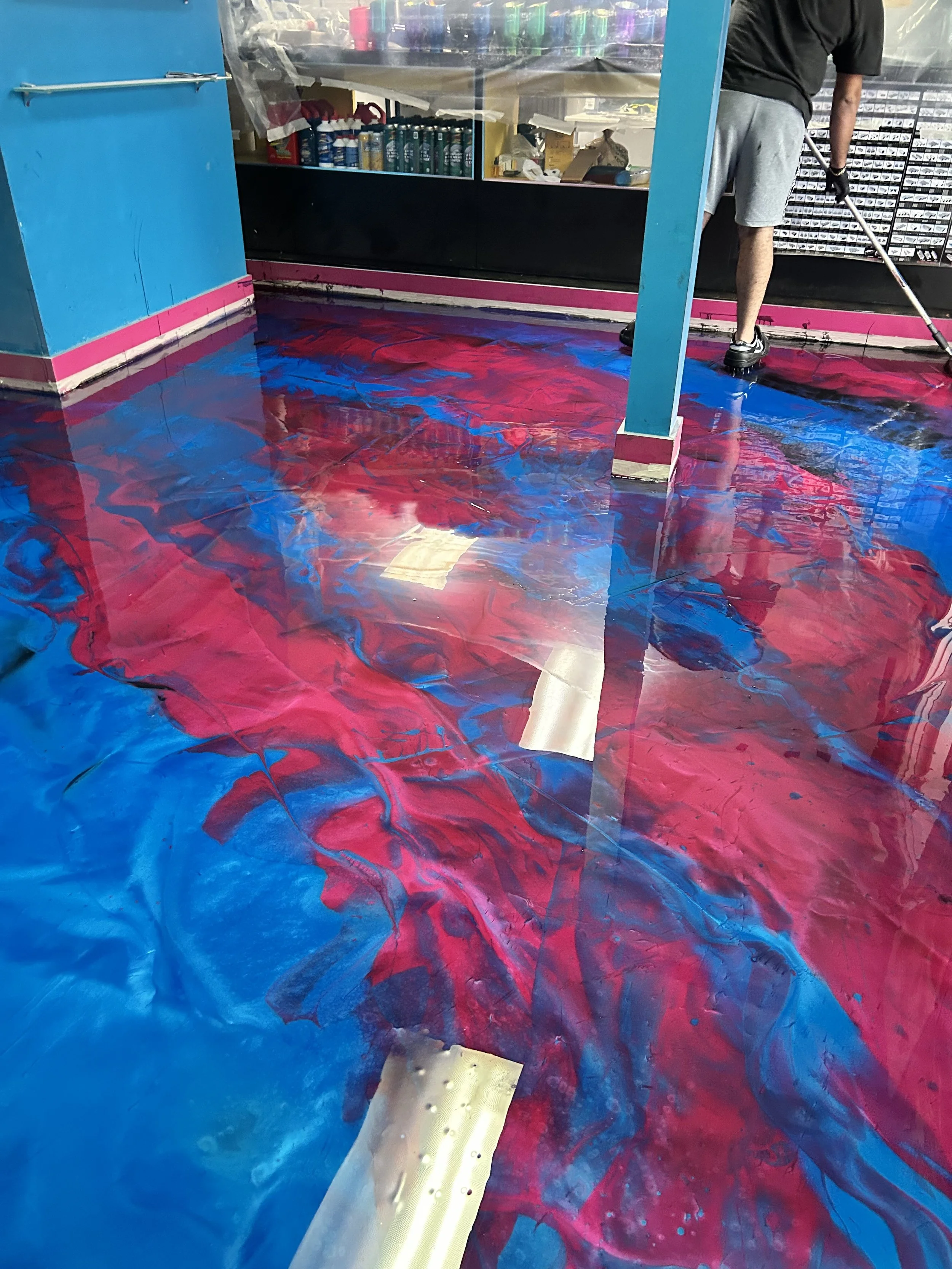 A person is cleaning a colorful floor with blue and pink swirling patterns inside a store or shop. The person wears gray shorts and black shoes, and is wiping the floor near a blue counter with various items inside.