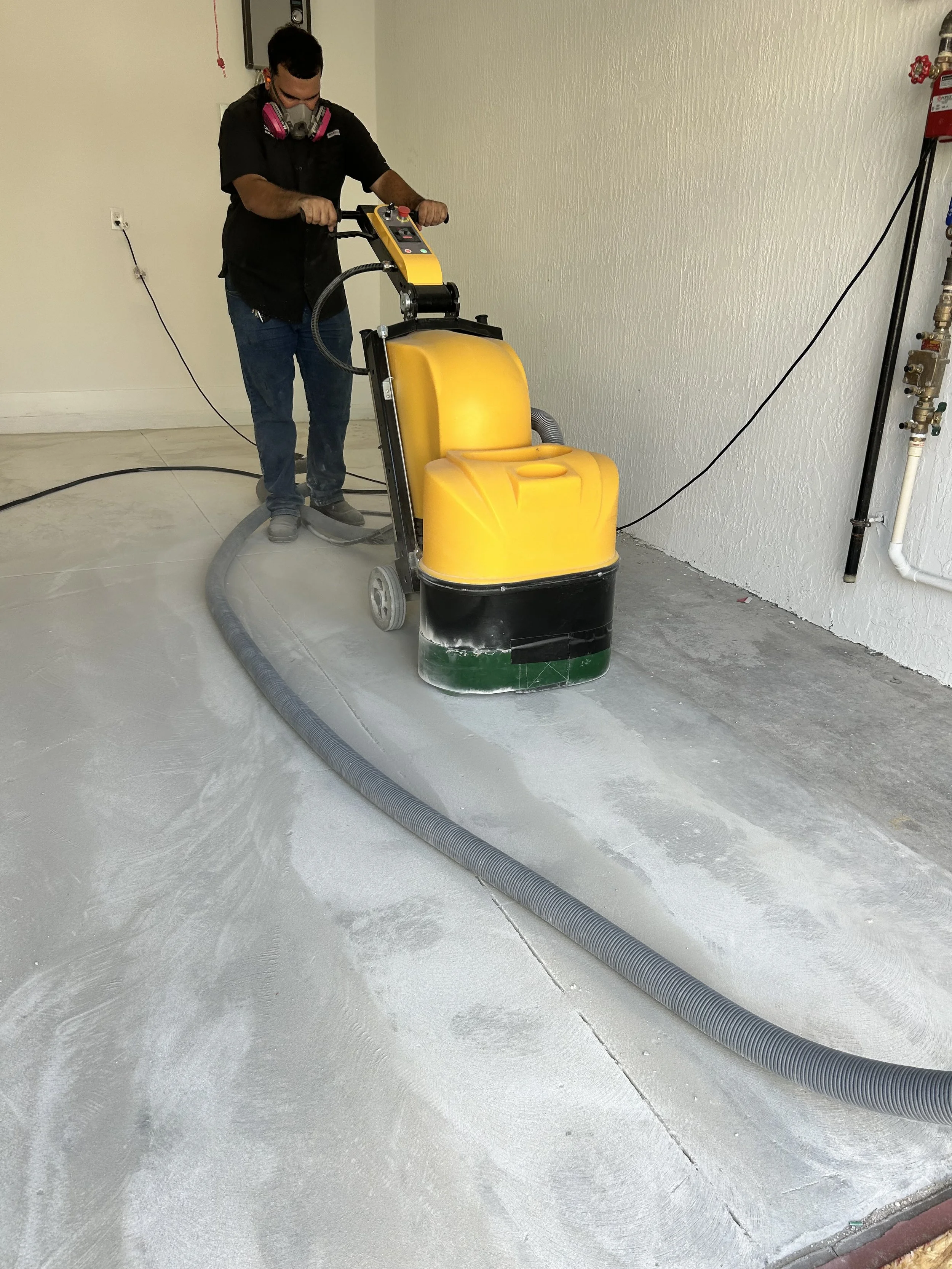 A person operating a yellow industrial floor cleaning machine, removing dust and debris from a concrete floor in a room with plain white walls.