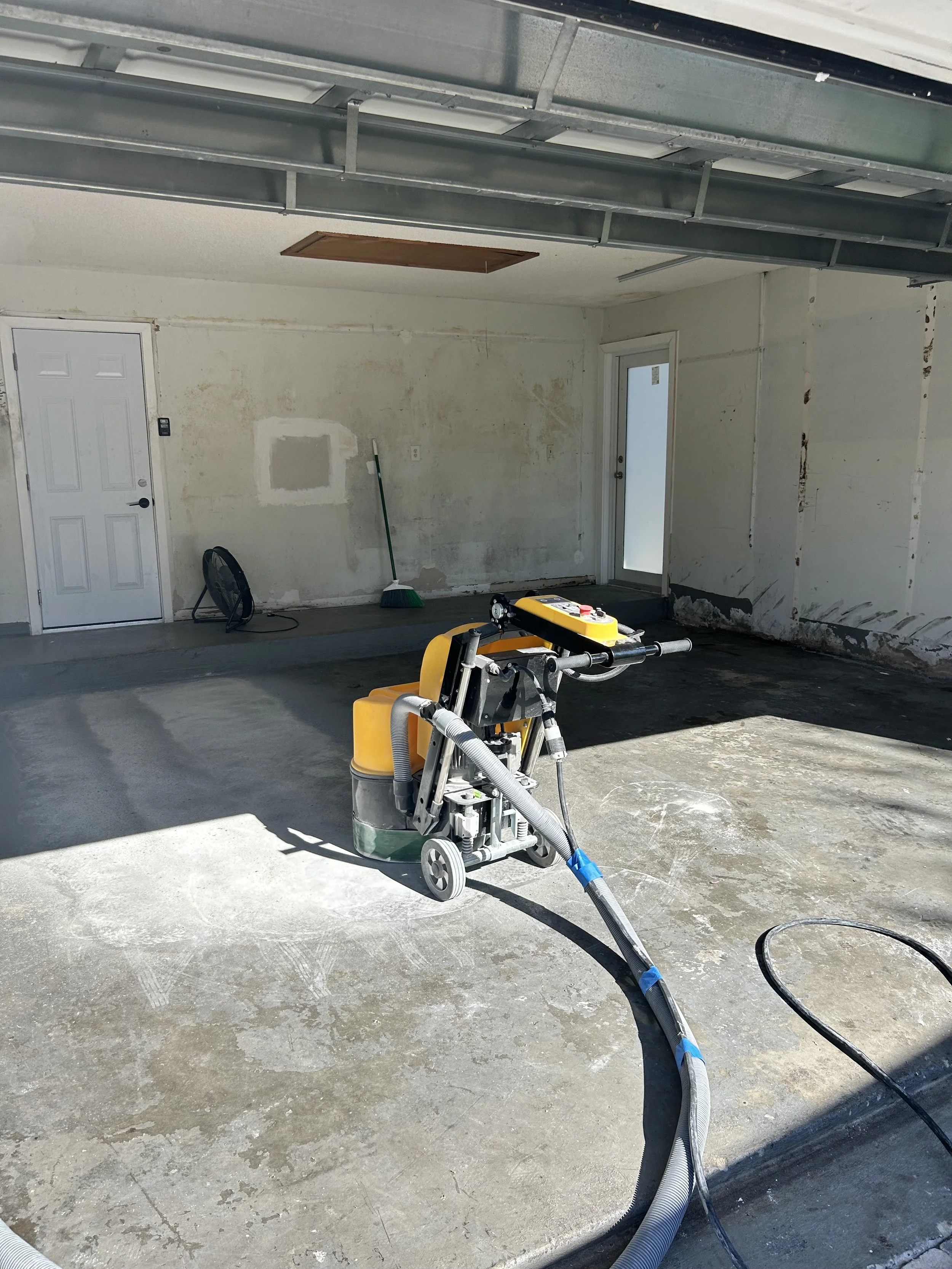 Garage under renovation with a concrete floor, a floor grinder machine, and cleaning tools like a broom and a fan, with unfinished walls and a partially open garage door.