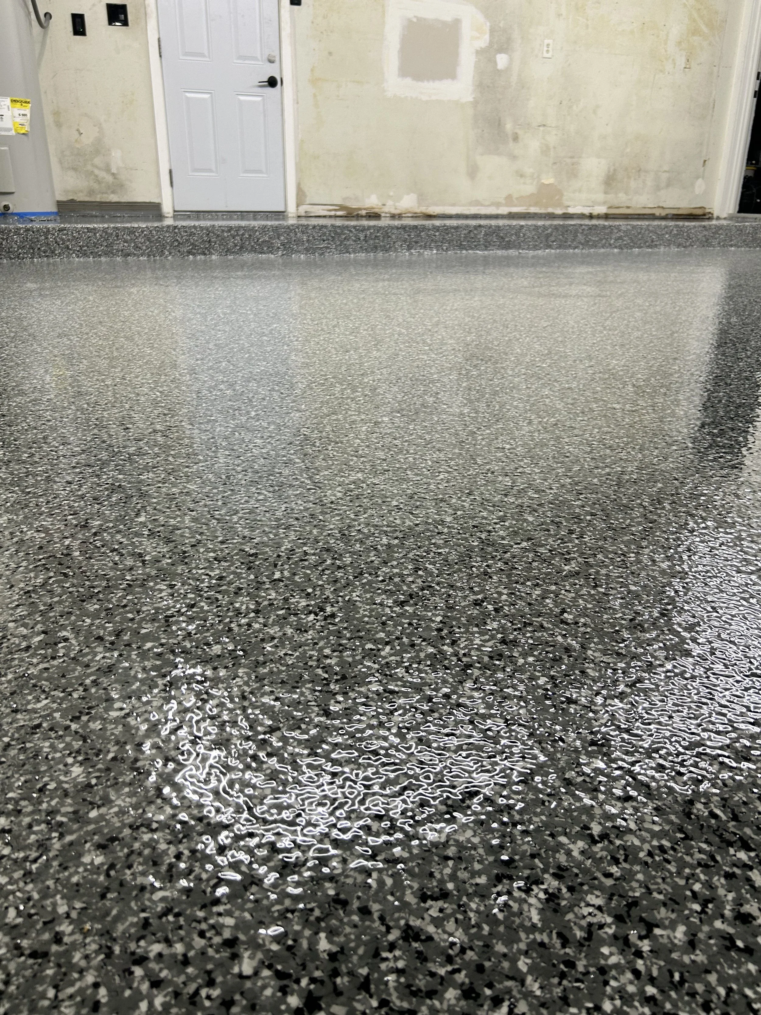 Polished speckled gray epoxy garage floor with a slight reflection. In the background, a white door and unfinished wall with patches of drywall and a small section of damaged wall are visible.
