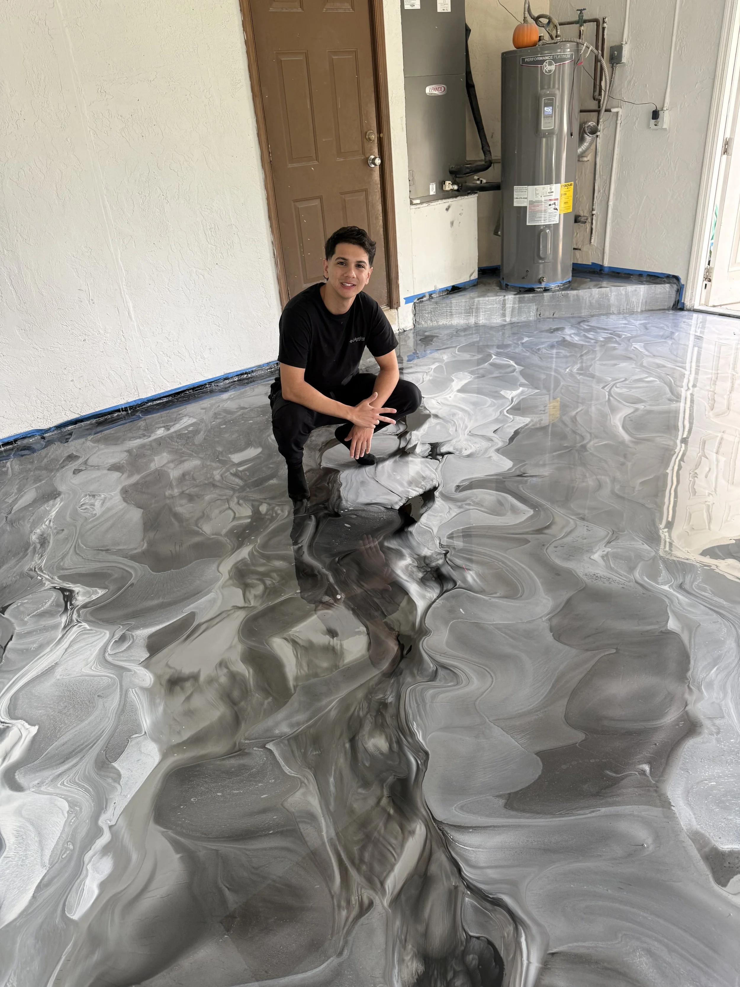 A young man squatting on a shiny, abstract-patterned concrete floor in a garage or utility room, with a water heater and electrical panel in the background.
