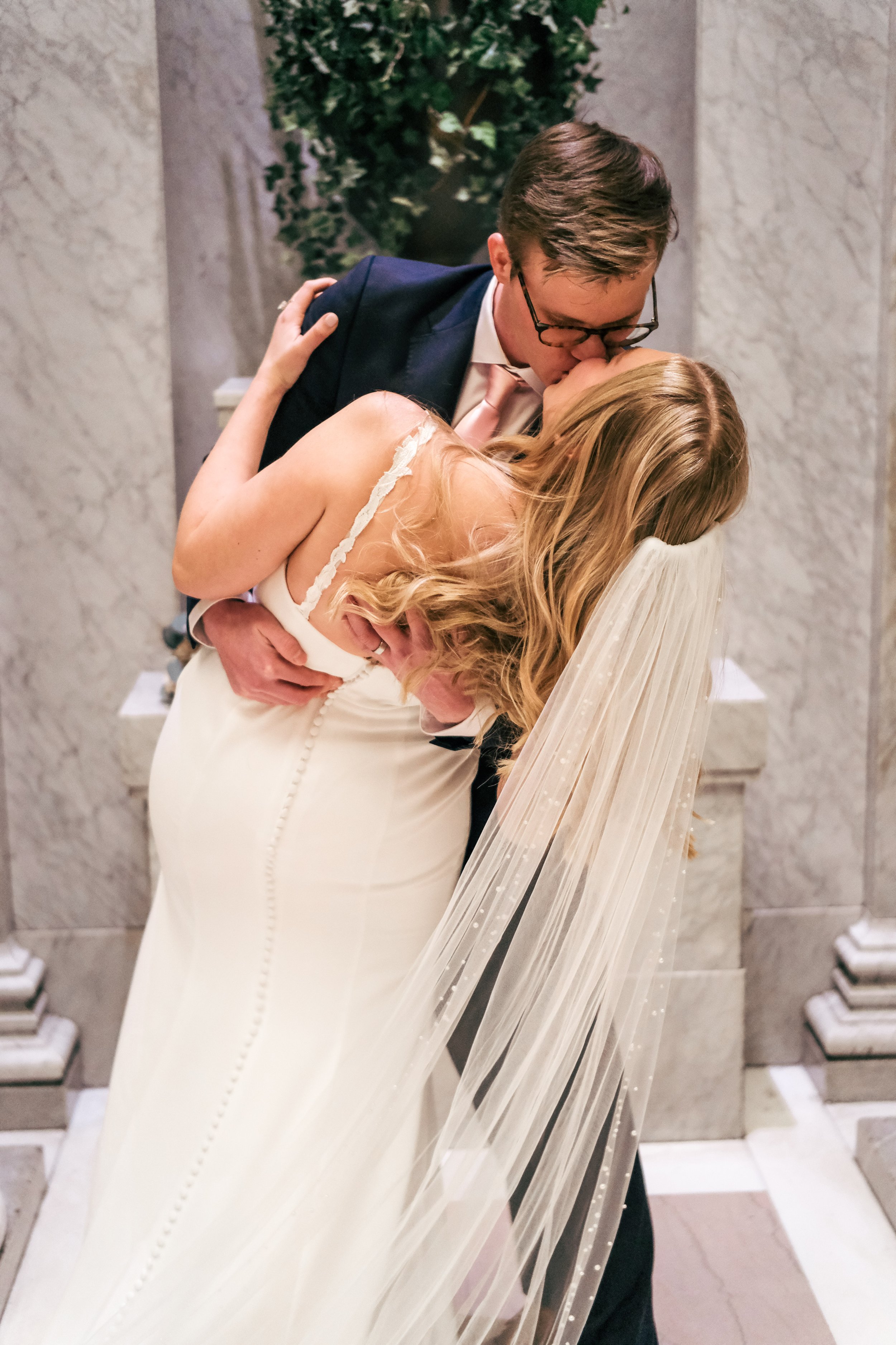 A couple shares a kiss in an elevator or hallway with marble walls. The woman wears a white dress with a veil, and the man wears glasses and a dark suit.
