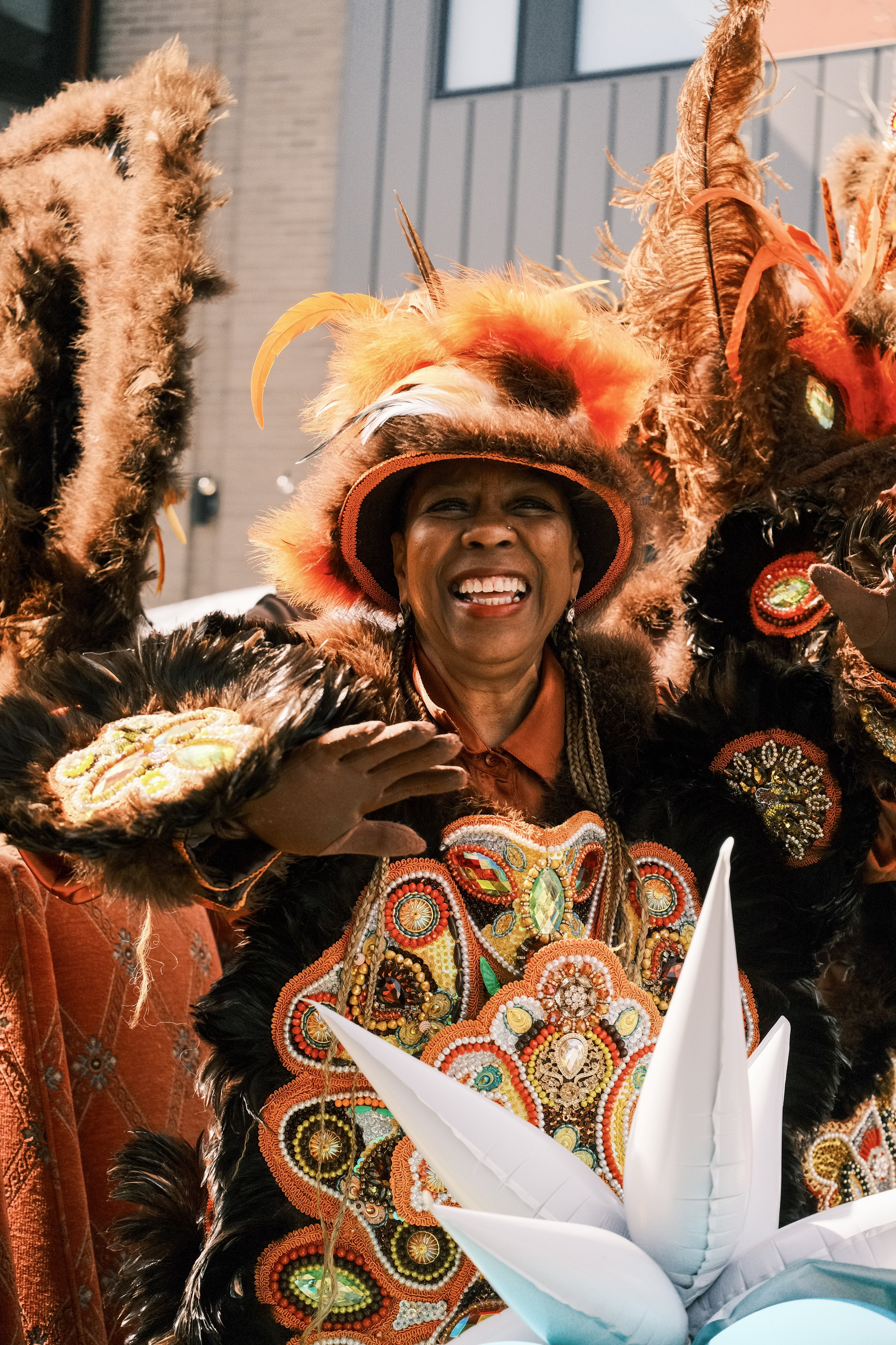 A woman in a vibrant, elaborate costume adorned with colorful beads, embroidery, feathers, and sequins, smiling and waving at a celebration.