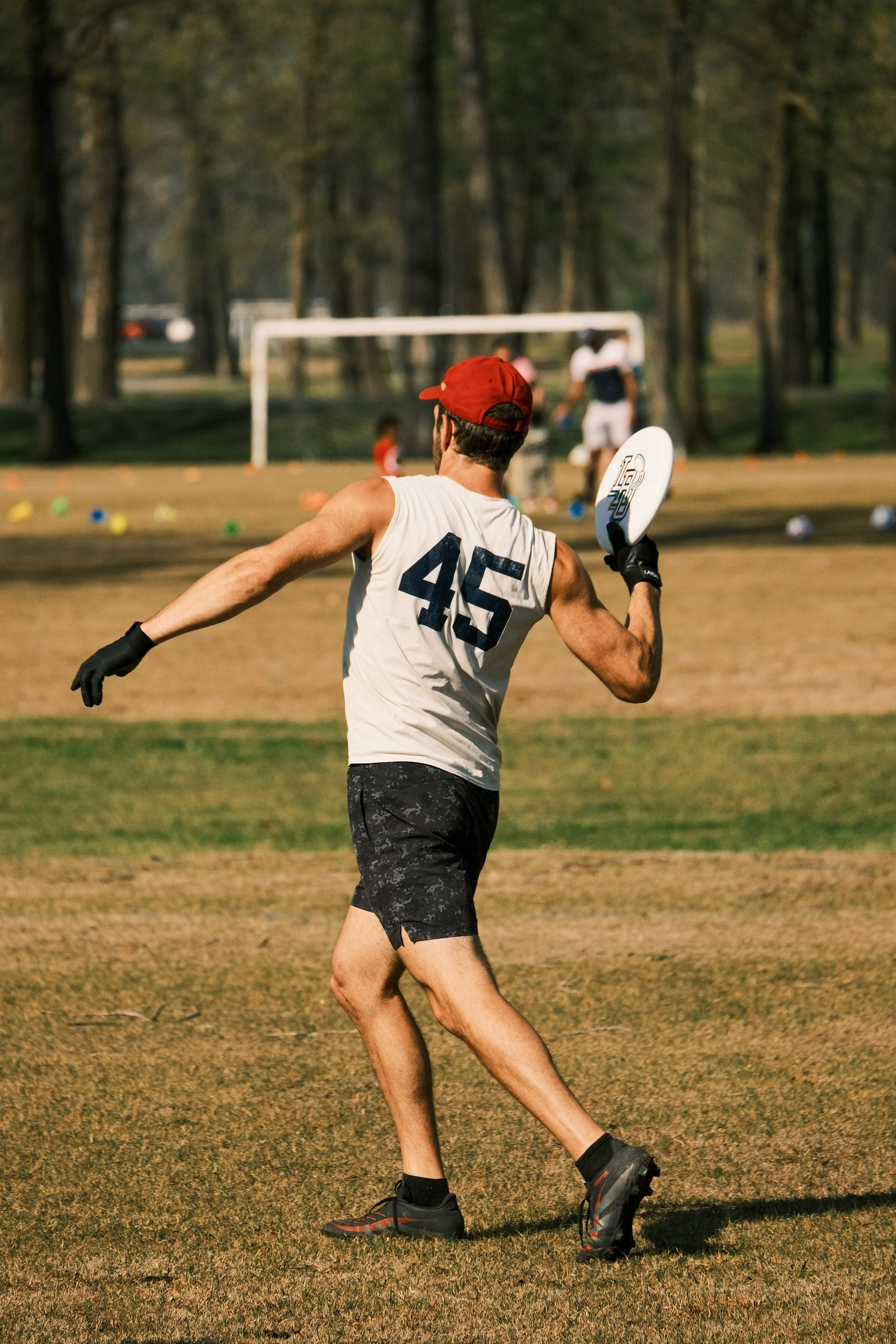A man in athletic clothing and a red cap running on a field with a frisbee in his hand, with children and a soccer goal in the background.