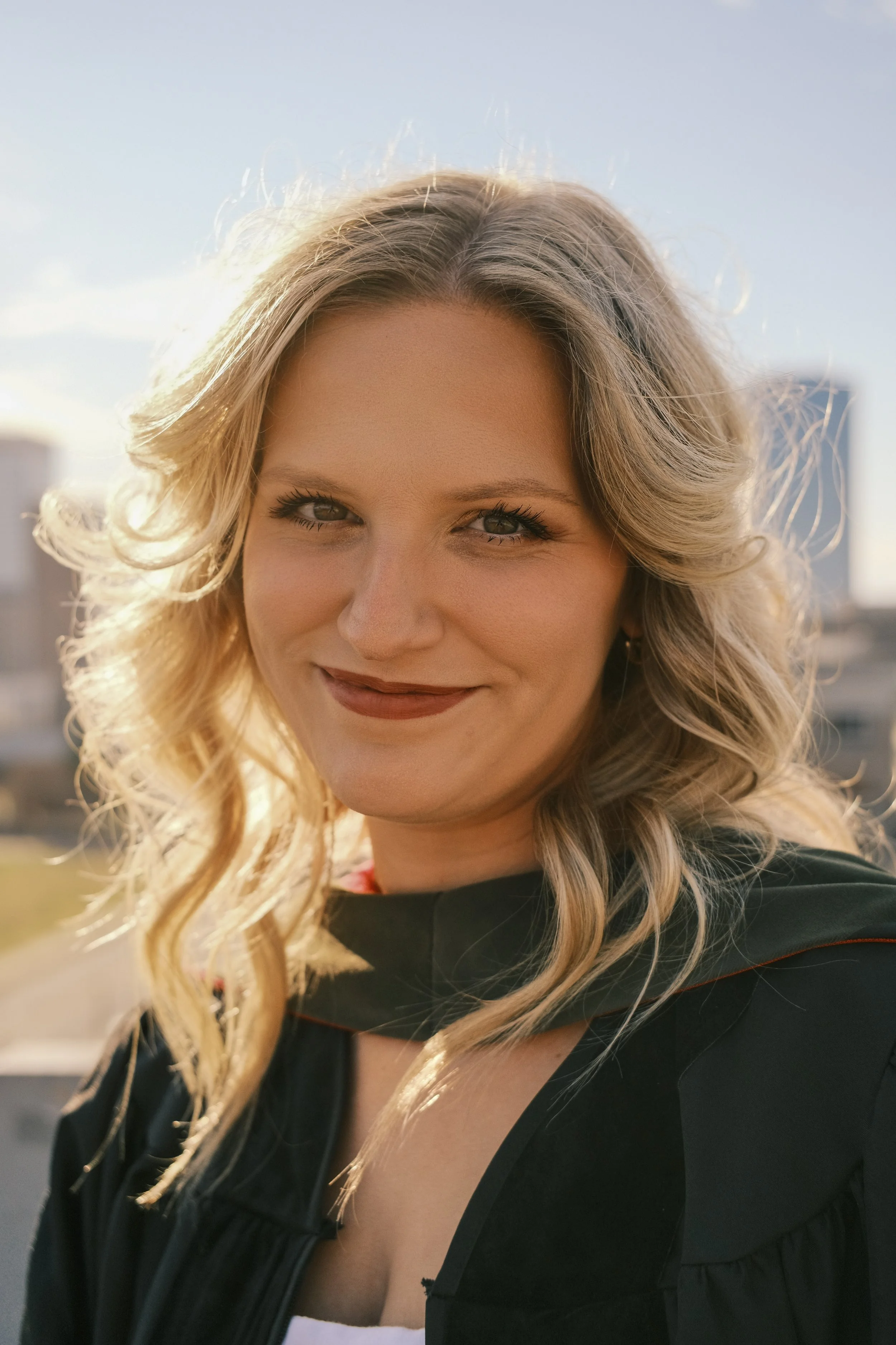 A woman with blonde, curly hair smiling outdoors in natural sunlight, wearing a black jacket over a white top.