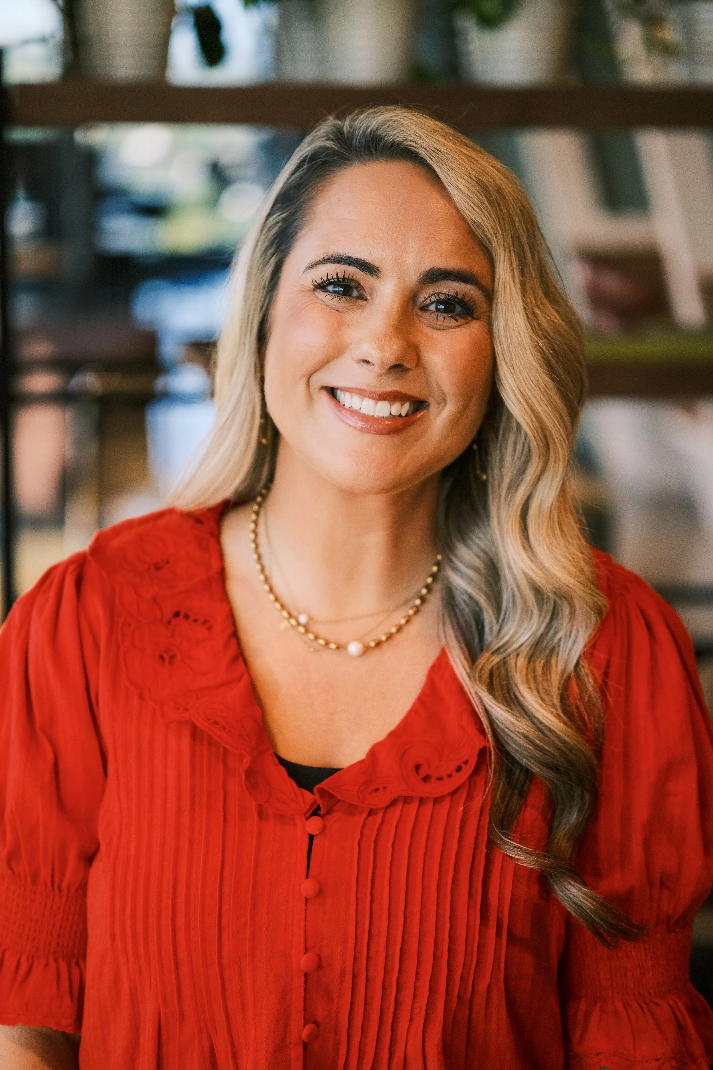 A woman with long wavy blonde hair smiling, wearing a red blouse with lace detailing and jewelry, standing indoors with a blurred bookshelf in the background.
