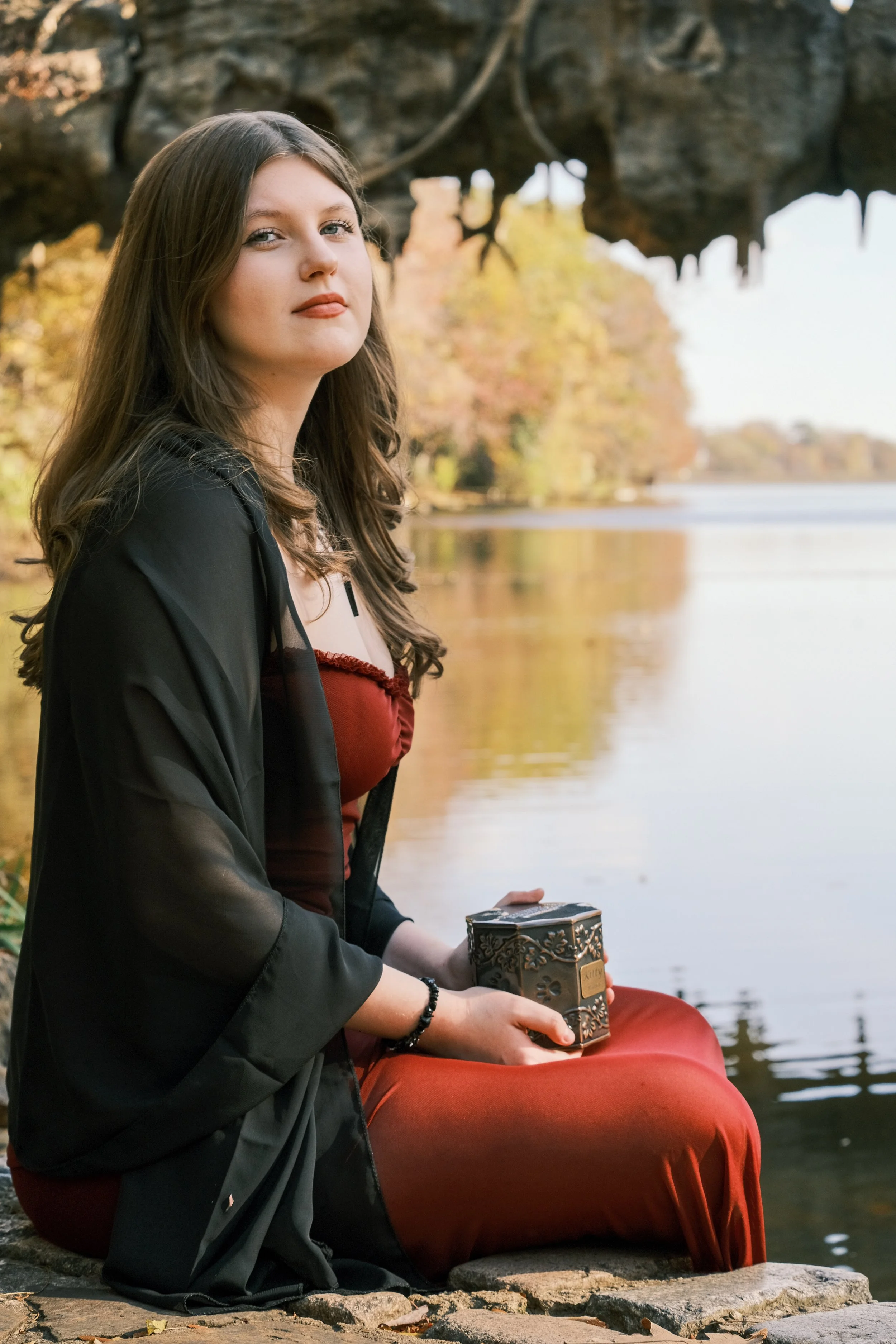 A young woman with long brown hair sitting on rocks by a lake, holding a decorative box, wearing a red dress and black sheer shawl, with autumn trees in the background.