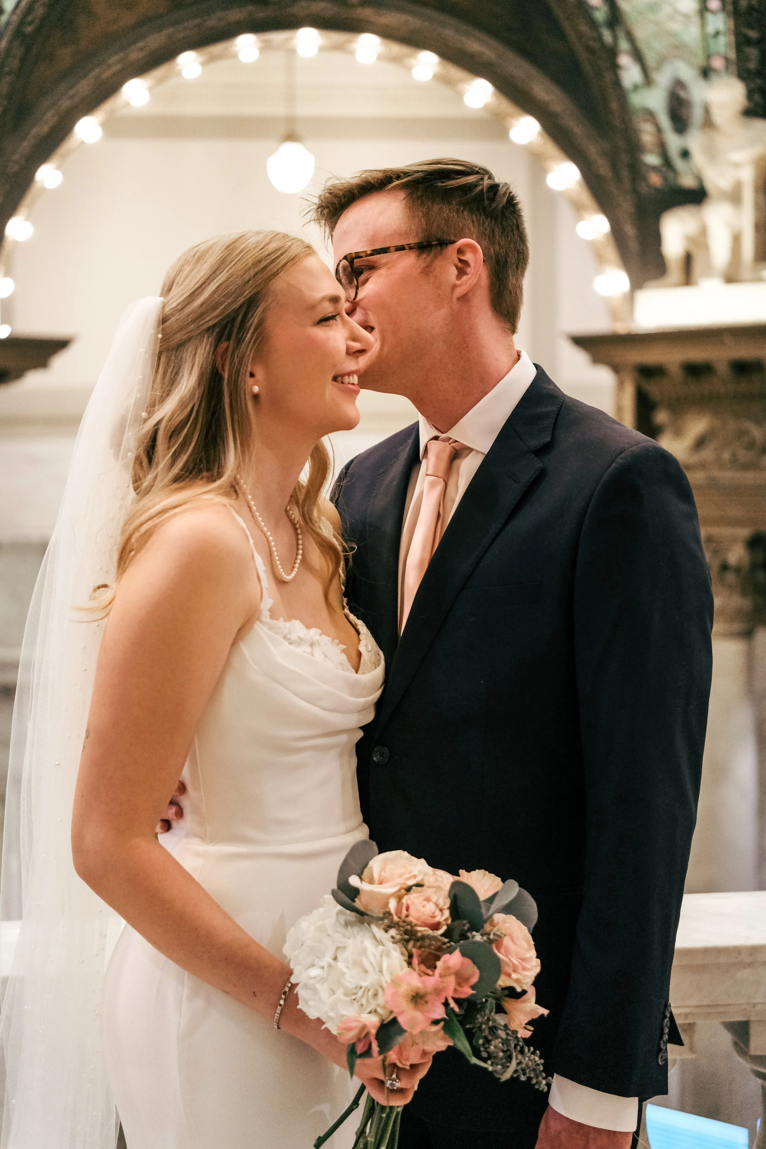 A bride and groom sharing a kiss during their wedding ceremony, with the bride holding a bouquet of pink and white flowers, and the groom wearing a navy blue suit with a light pink tie.