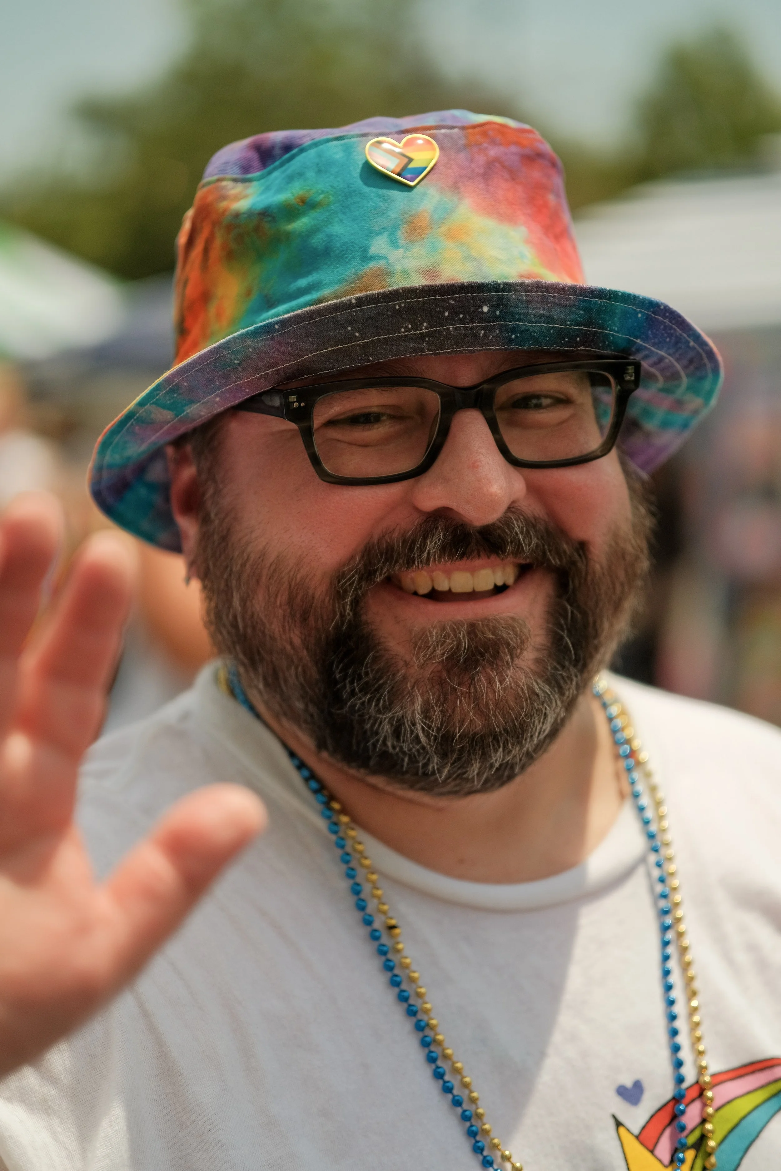 A man wearing glasses and a colorful tie-dye bucket hat with a rainbow rainbow heart pin, smiling and waving, with beads around his neck, in a crowded outdoor setting.