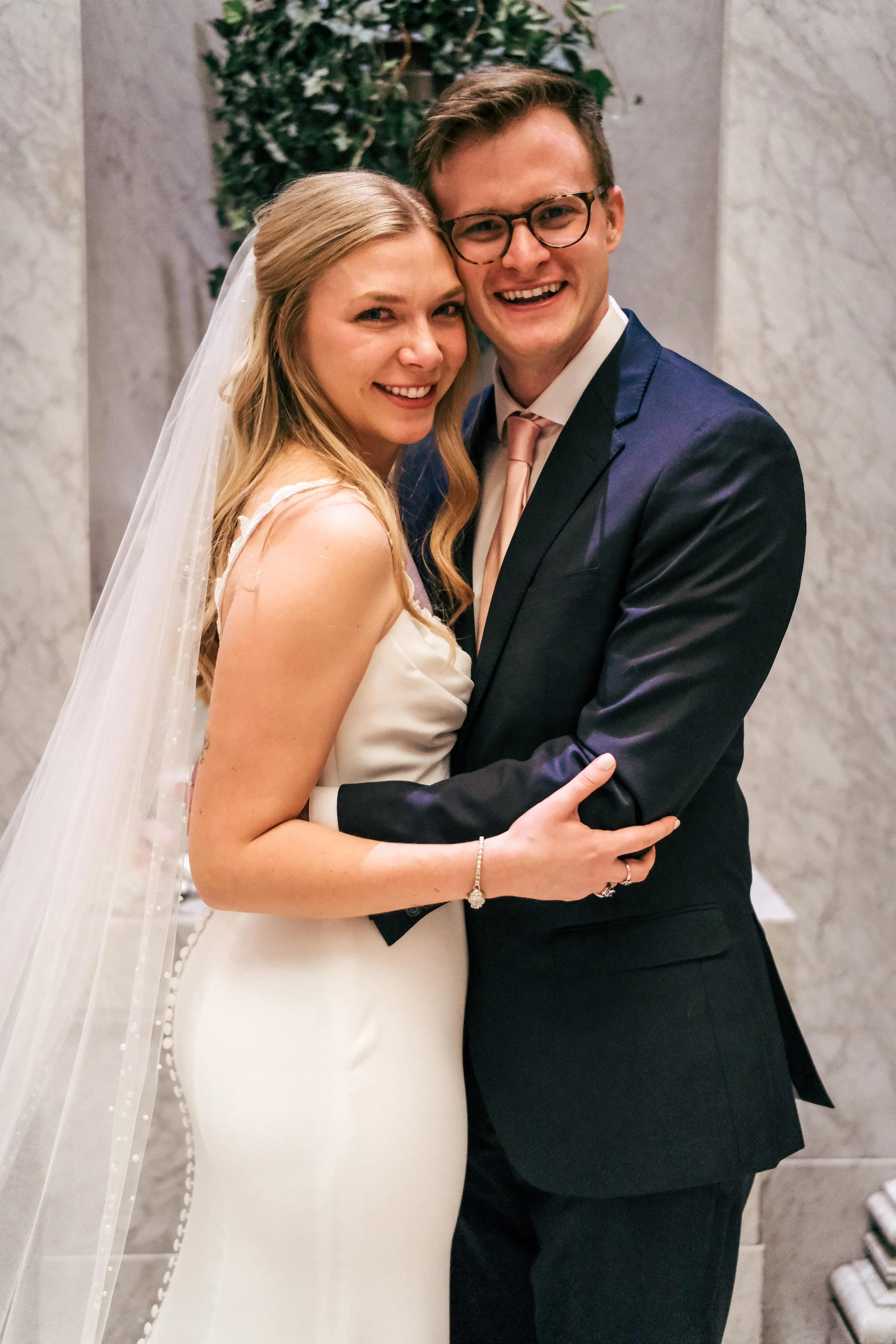 A newlywed couple smiling and embracing each other; the bride is wearing a white wedding dress with a veil, and the groom is in a dark blue suit with a pink tie; they are standing indoors against a marble wall with greenery in the background.