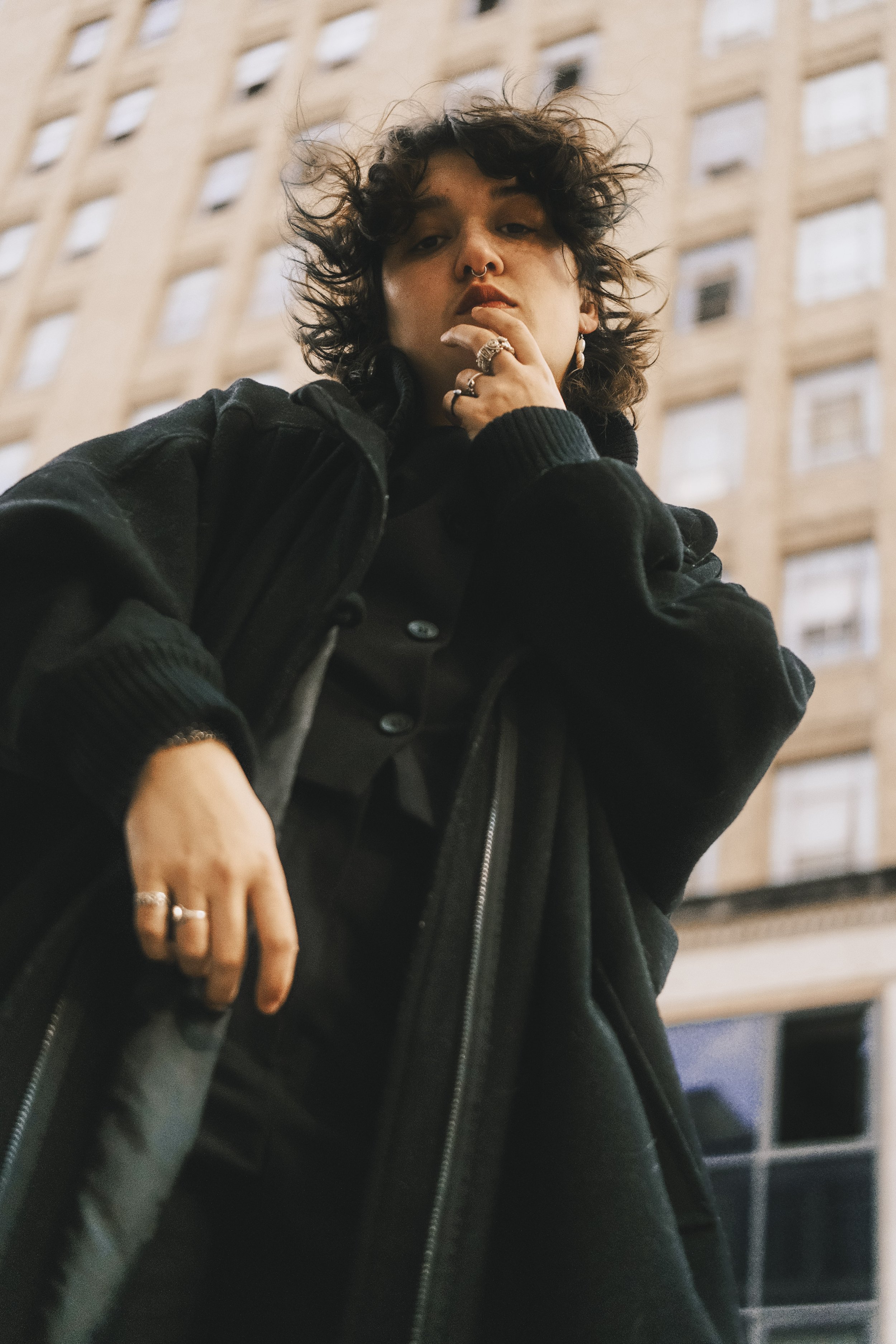 A young person with curly hair, multiple rings, and a septum piercing posing in front of a tall building, wearing a black jacket.