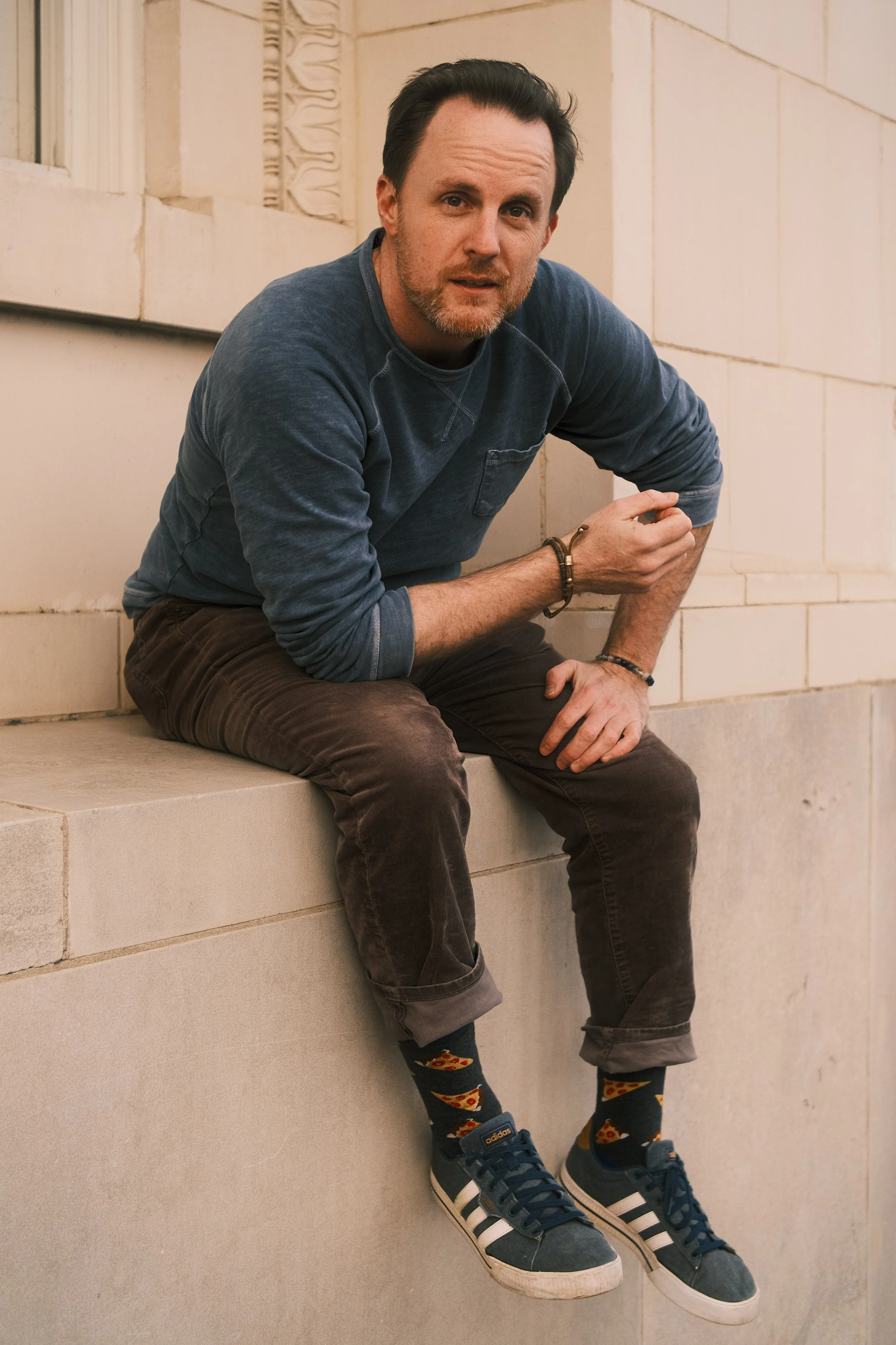 A man with dark hair and a beard, wearing a blue sweatshirt, brown pants, colorful pizza-themed socks, and blue Adidas sneakers, sitting on a ledge with a brick wall in the background.