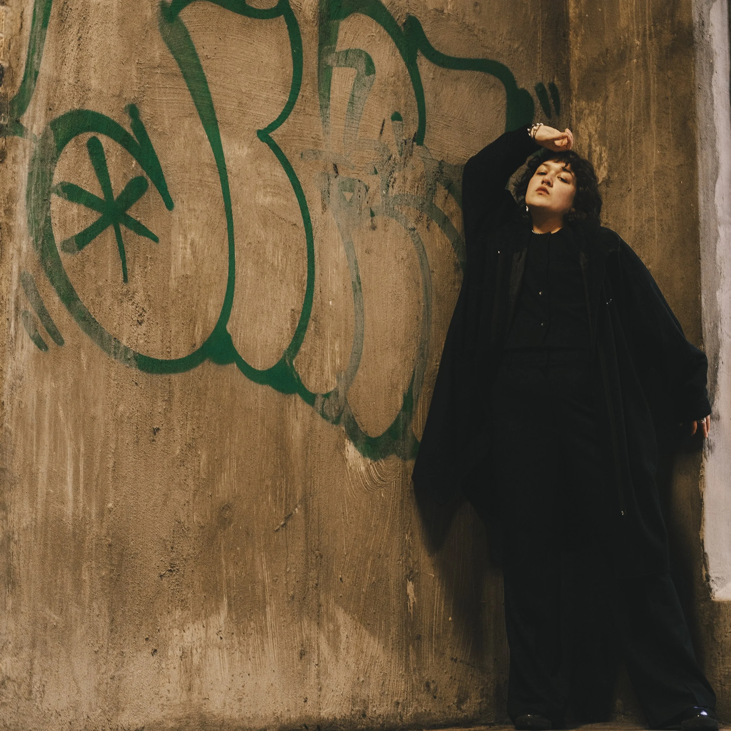 A person with dark curly hair wearing all black clothing, leaning against a concrete wall with graffiti in a dimly lit environment.