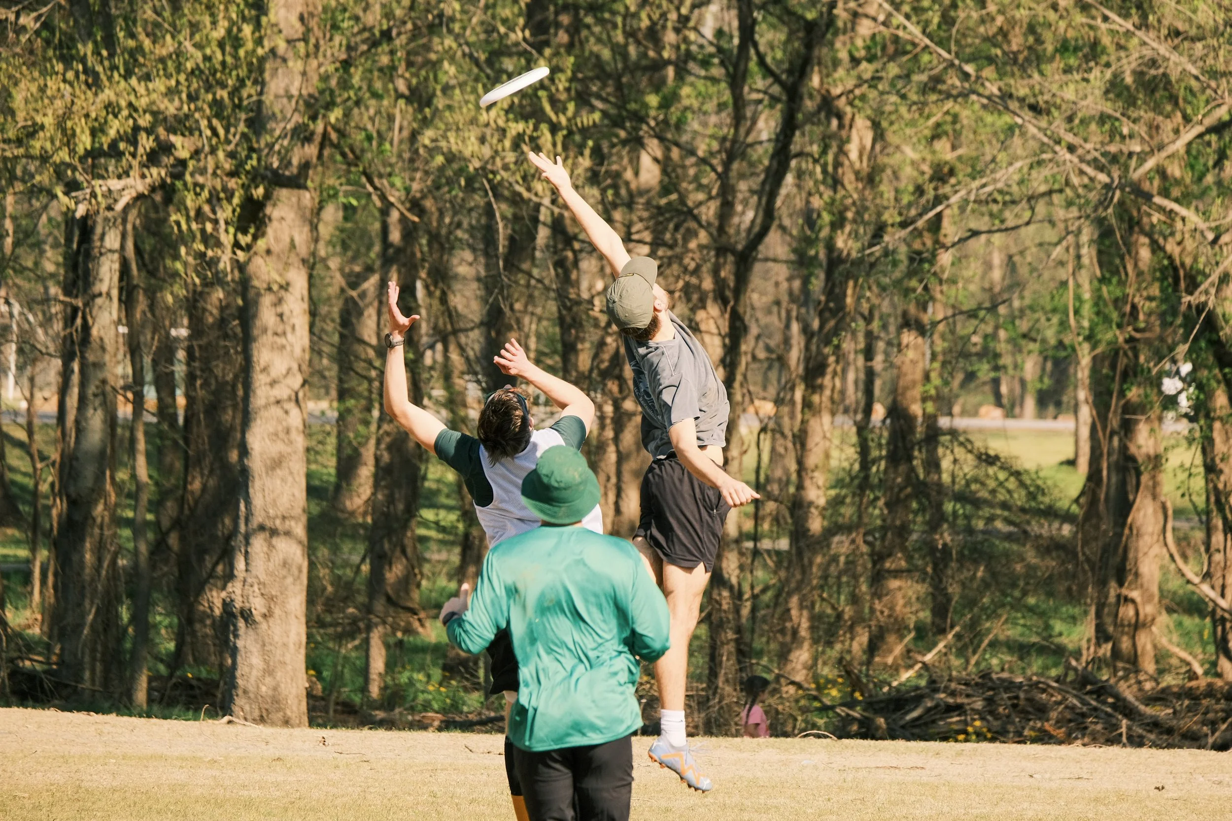 Two young boys playing disc golf outdoors on a sunny day, with one boy in mid-air reaching to catch or throw a frisbee, set against a backdrop of trees.
