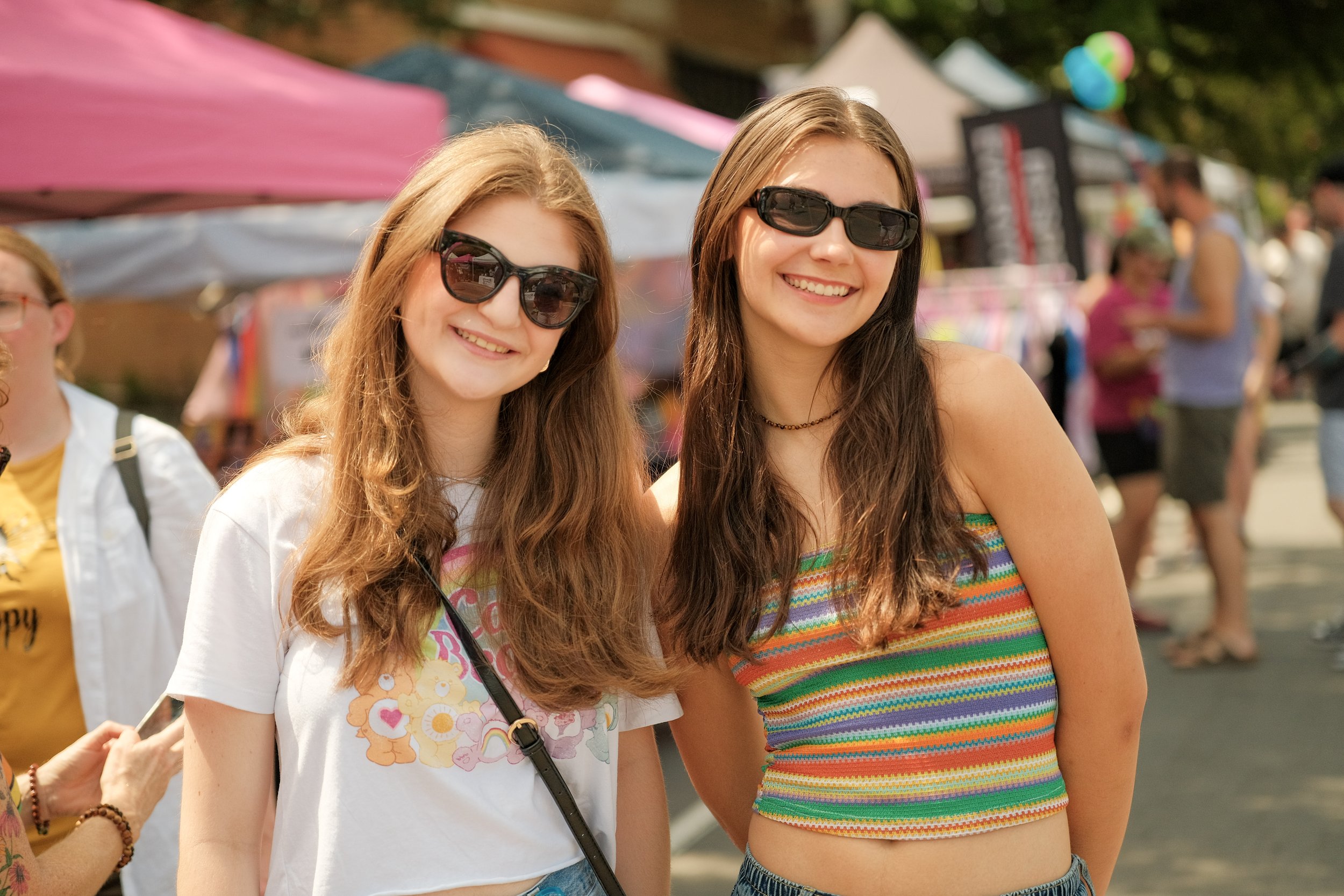 Two young women wearing sunglasses smiling at a fair or outdoor event with booths and balloons in the background.