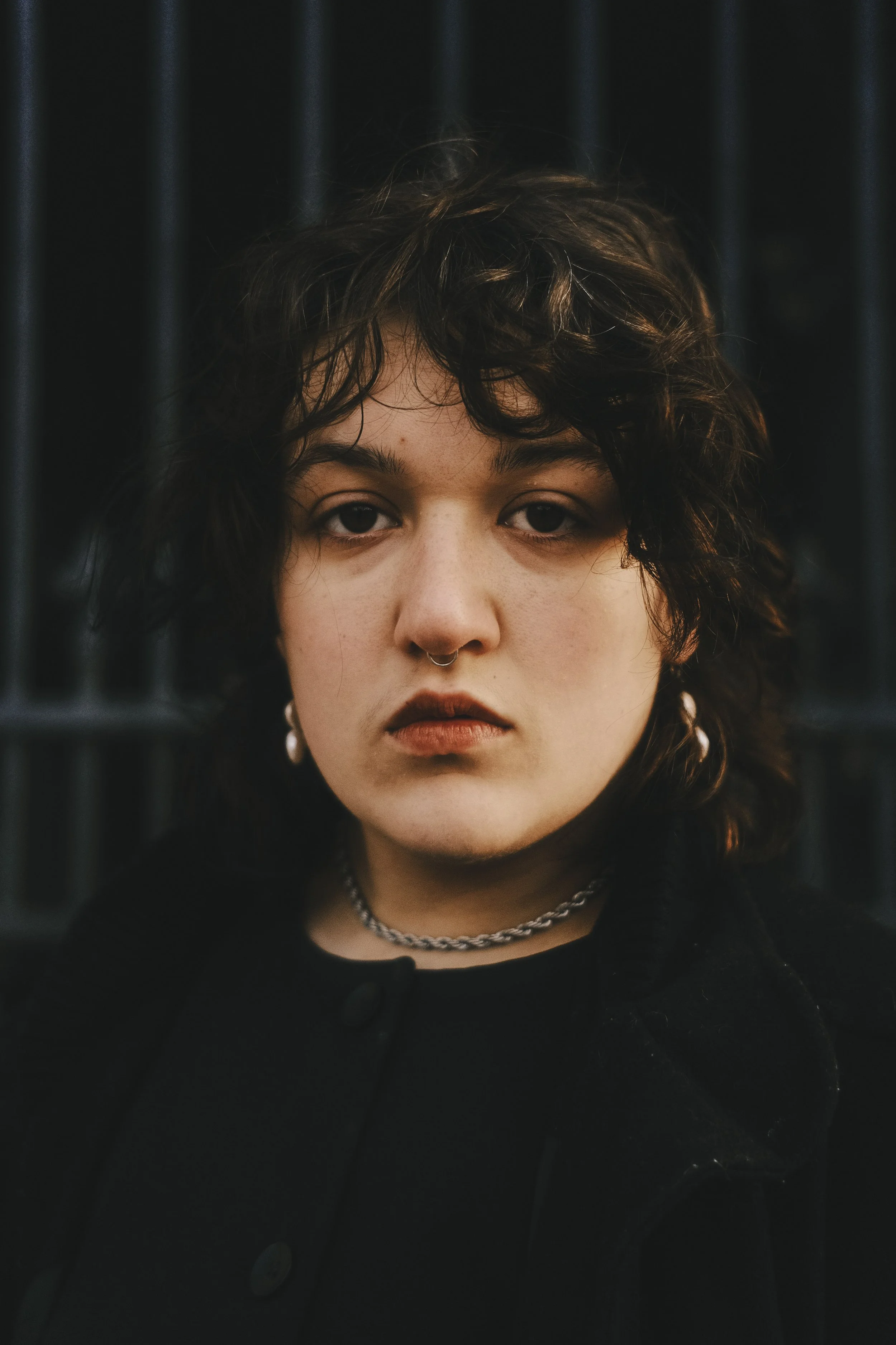 A young person with curly dark hair, wearing a black jacket and layered necklaces, standing in front of black bars, staring directly at the camera with a neutral or serious expression.